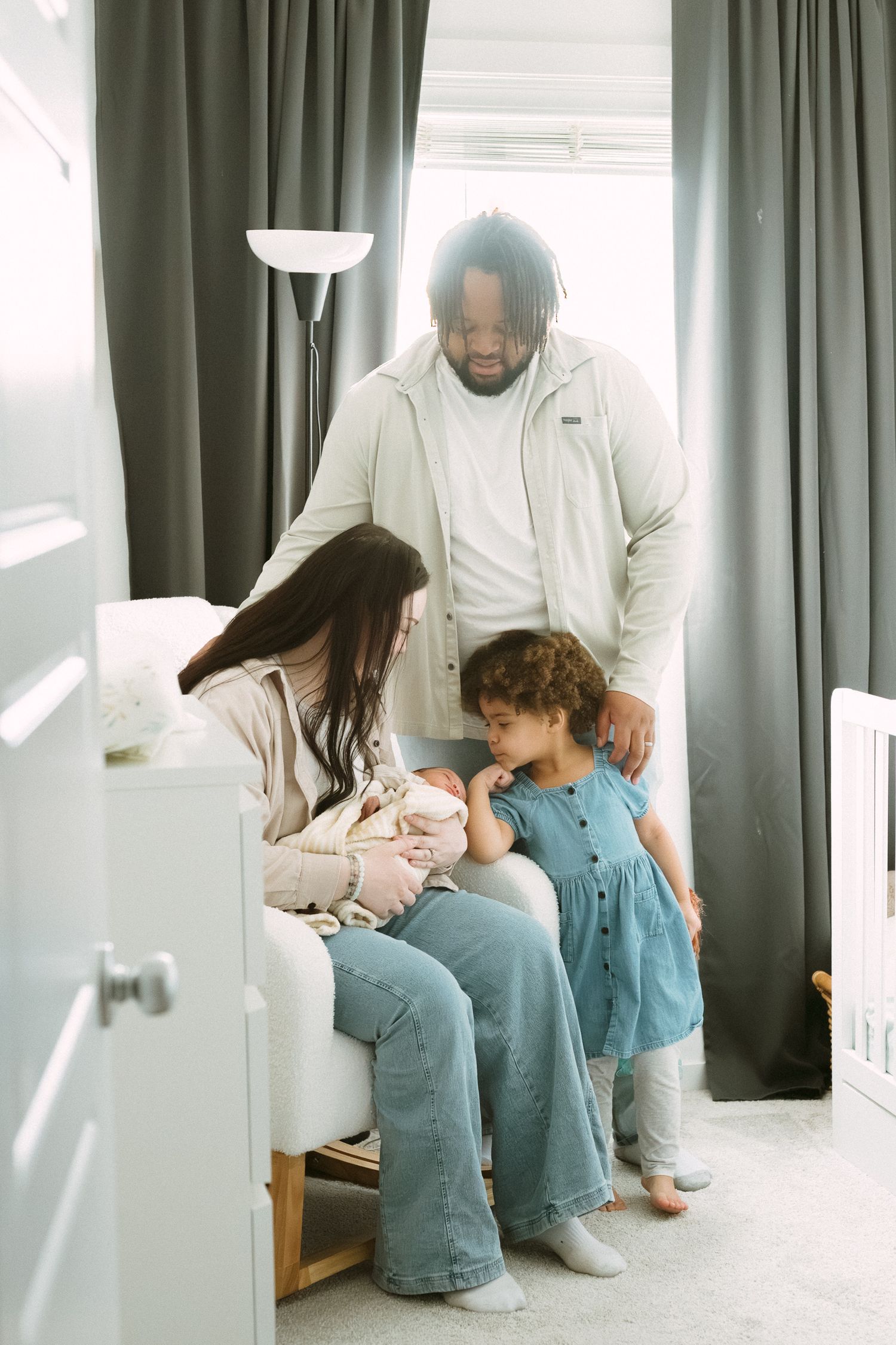 A multiracial family admires their newborn baby in a sunlit nursery, with mom seated and dad and toddler looking on.