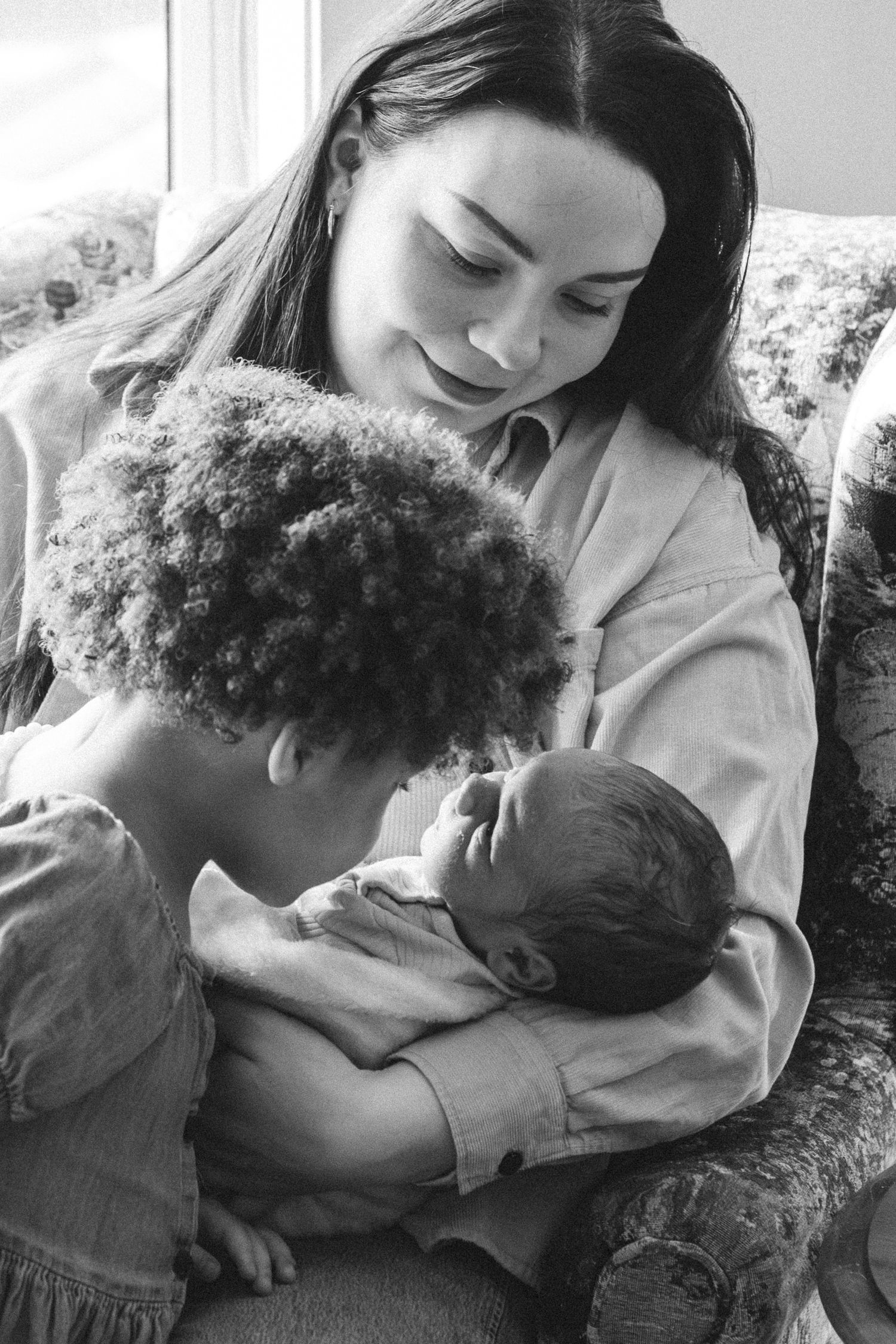 Black and white photo of a young mother sitting in a chair, tenderly holding a newborn while an older child looks on.
