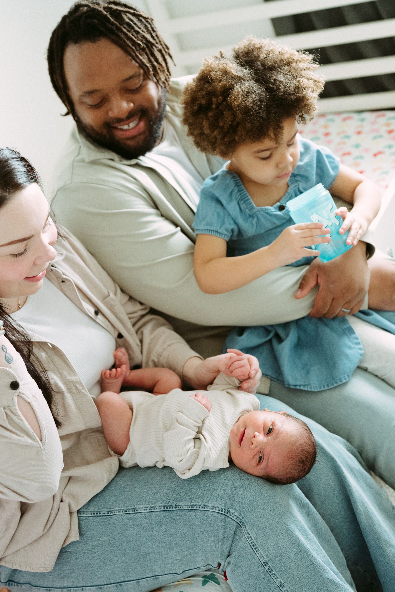 A smiling family with a newborn baby and toddler relaxing together on a bed in a cozy bedroom.