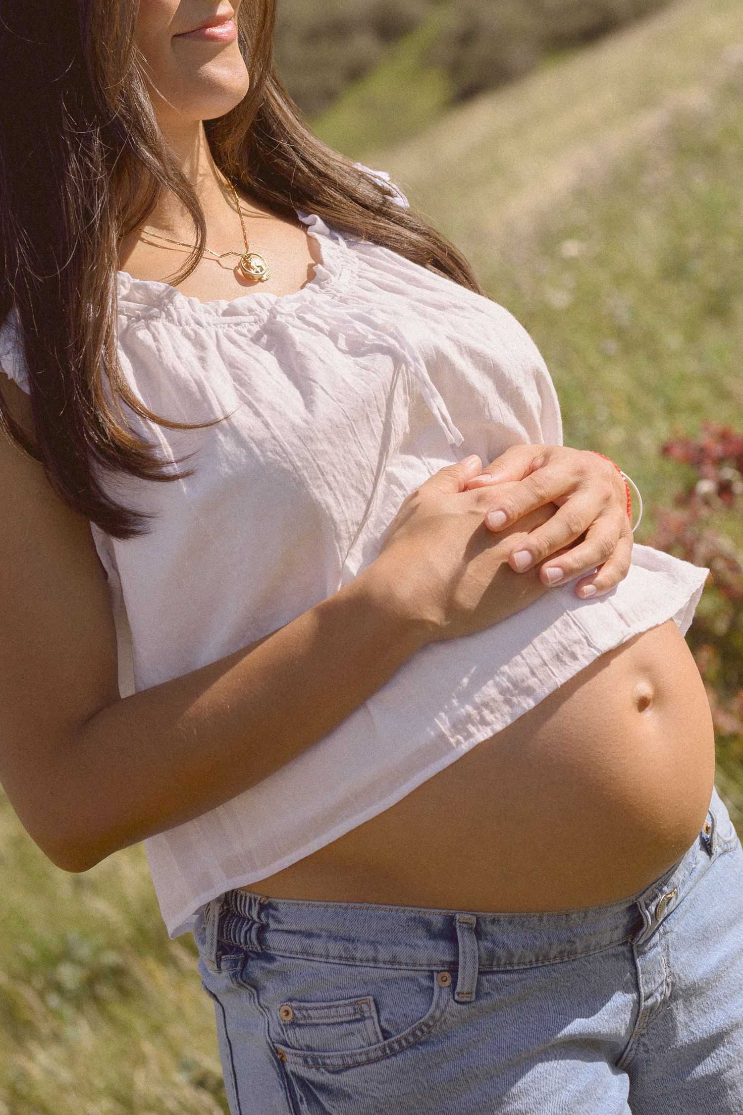 Close-up of a pregnant belly with hands resting gently on top in natural sunlight.