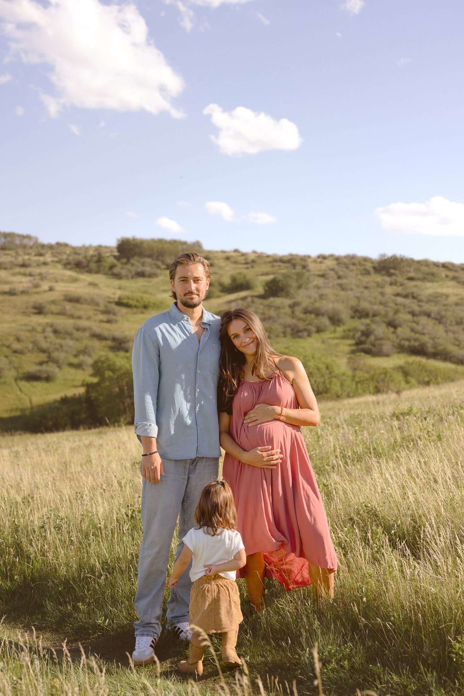 A family stands together in a sunlit meadow with rolling hills in the background.