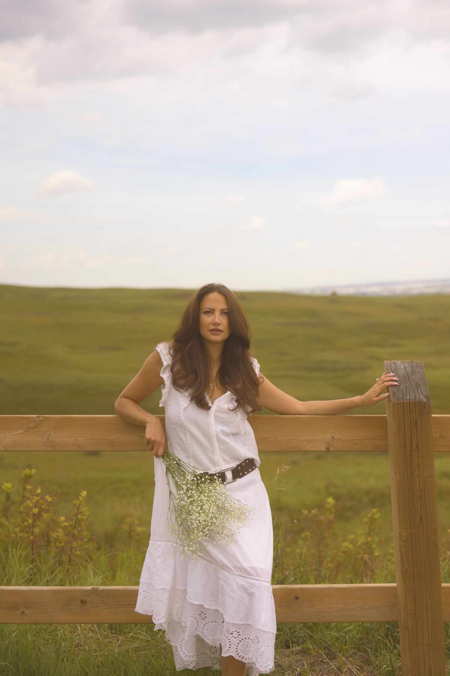 Figure in white dress holds onto rustic fence post in pastoral landscape.
