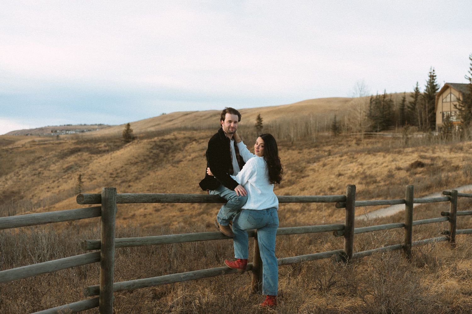 A couple shares a romantic moment along a rustic wooden fence in a scenic autumn landscape at sunset.