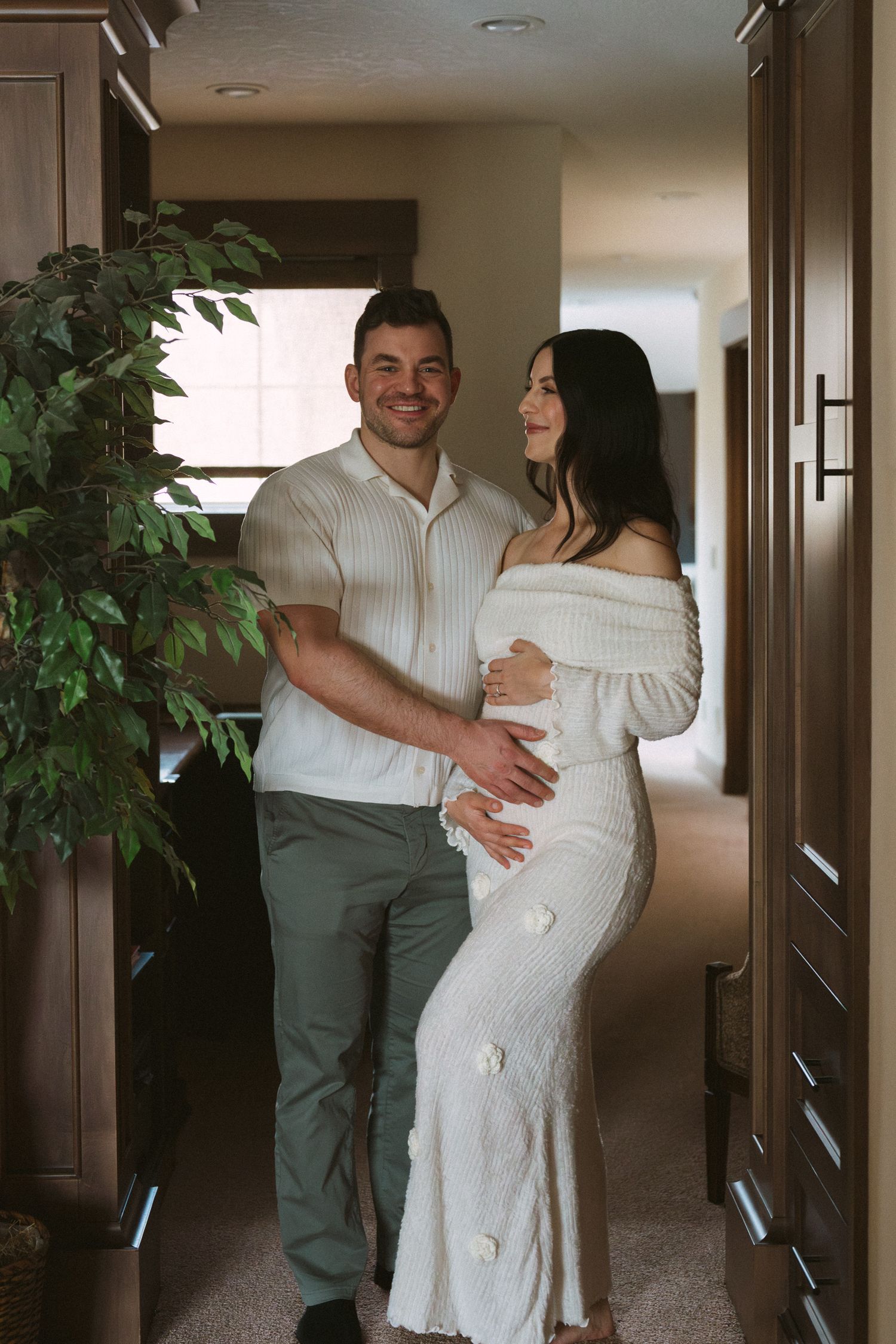 A couple poses indoors, man in white shirt embraces pregnant woman in white off-shoulder gown, smiling warmly.