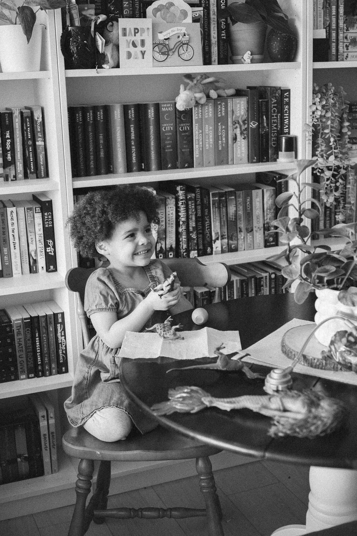 A young girl with curly hair sits at a wooden table smiling, surrounded by bookshelves filled with books.