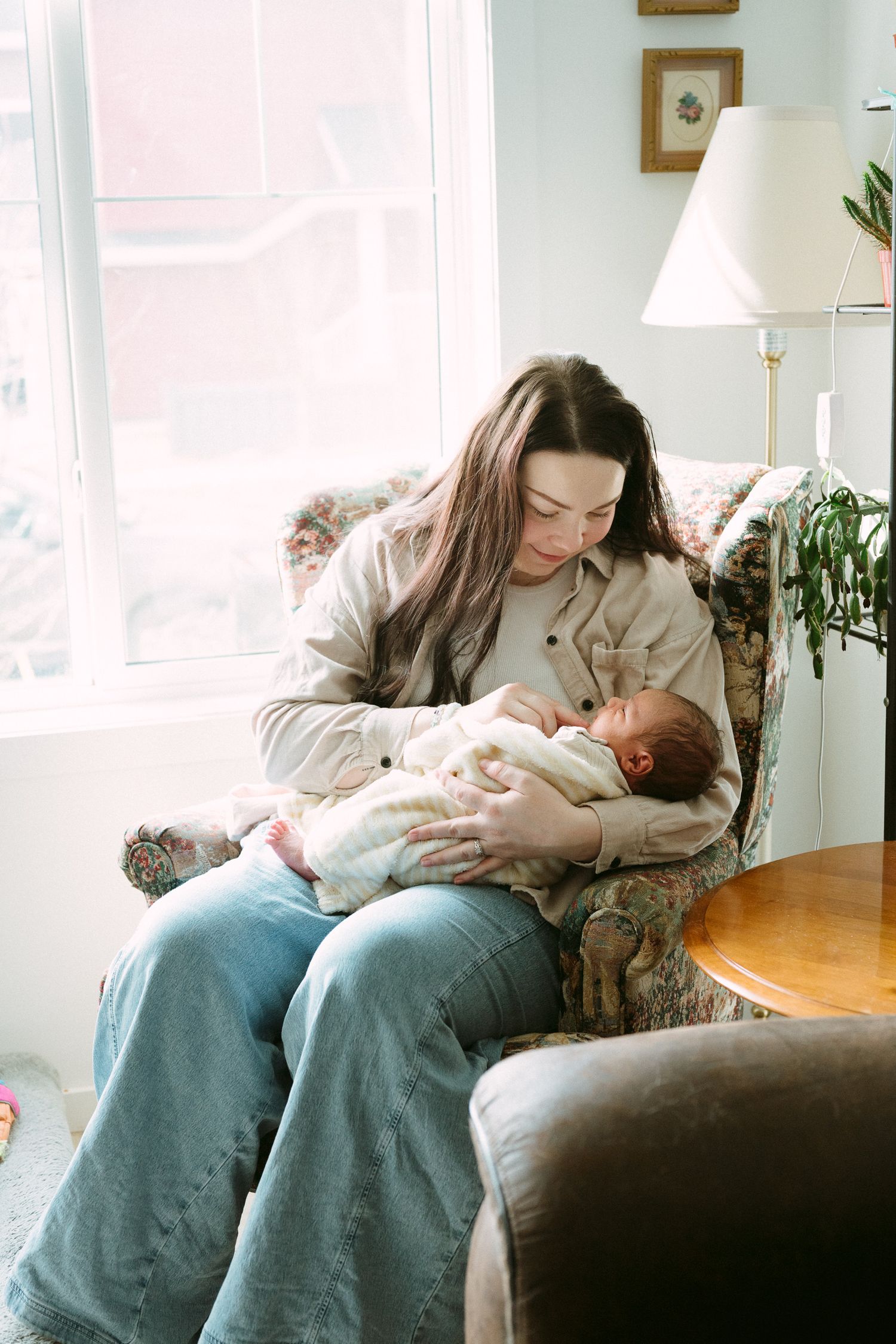 A young mother sits in a floral armchair by a window, tenderly cradling her newborn baby in soft natural light.