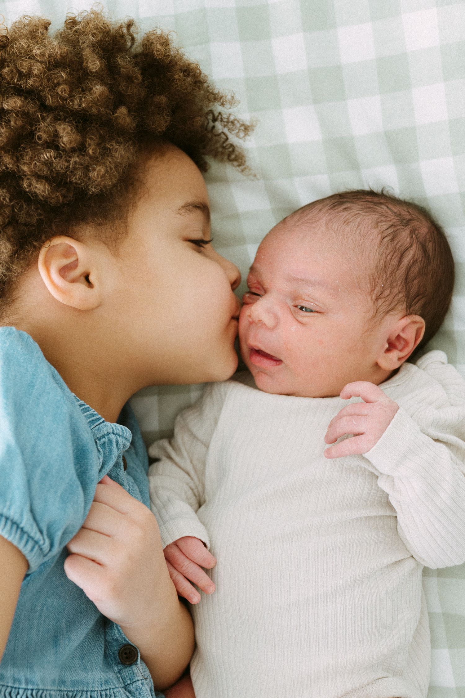 Young child with curly hair kissing a newborn baby lying on a green gingham blanket.