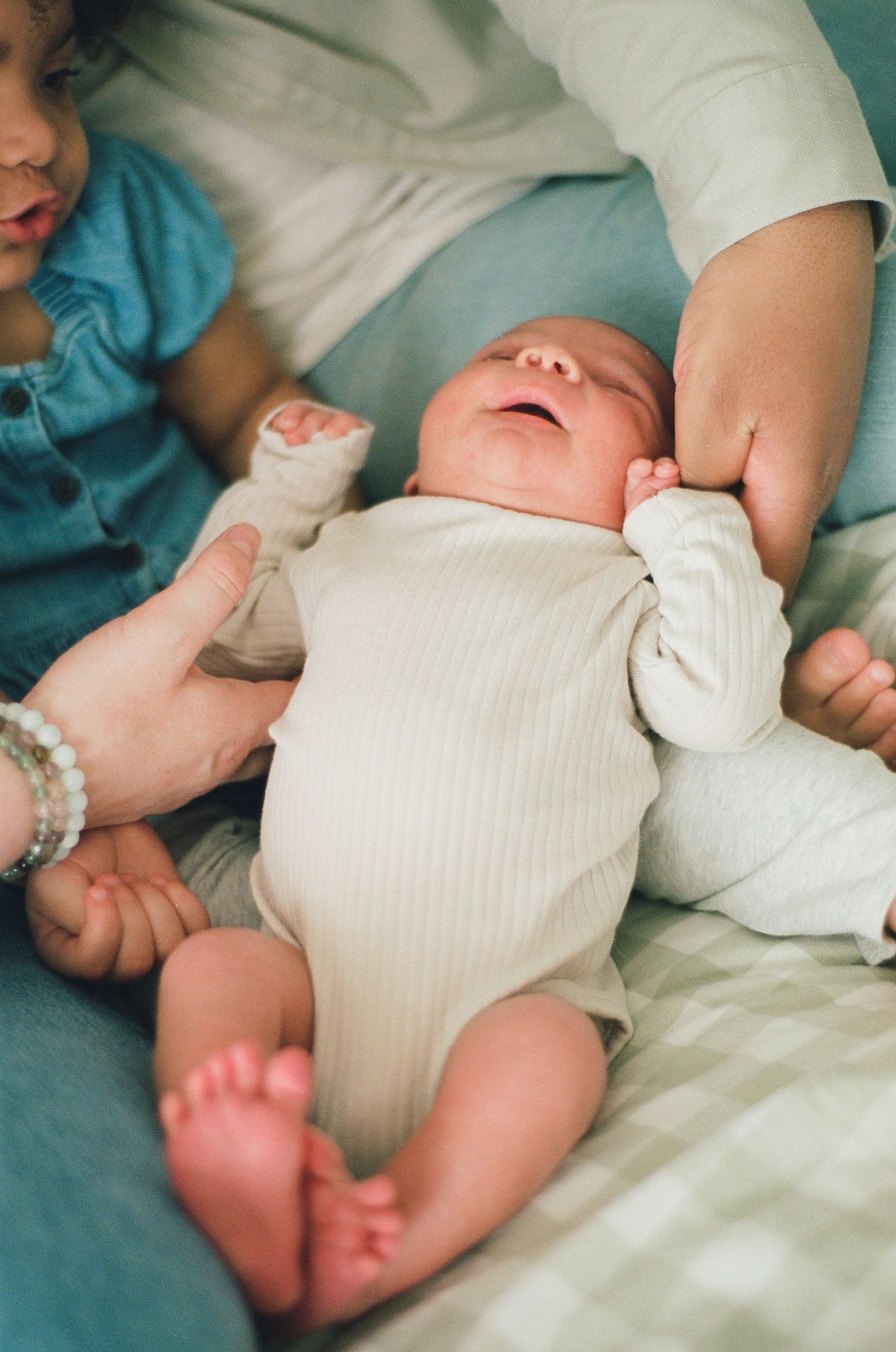 Newborn baby in white onesie lying on bed while older sibling and parent gently hold them.