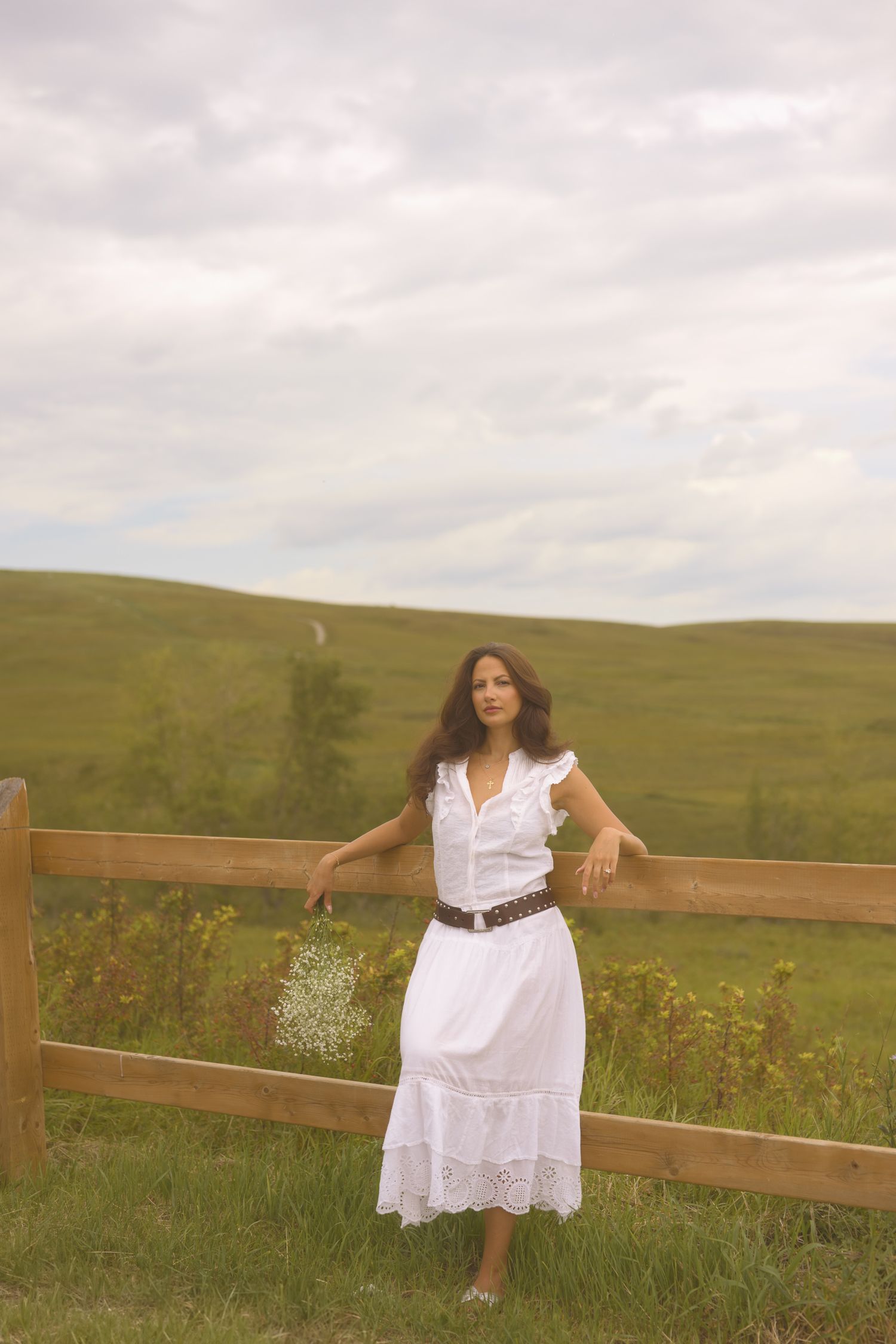 Person in white dress poses by wooden fence in rolling countryside meadow.