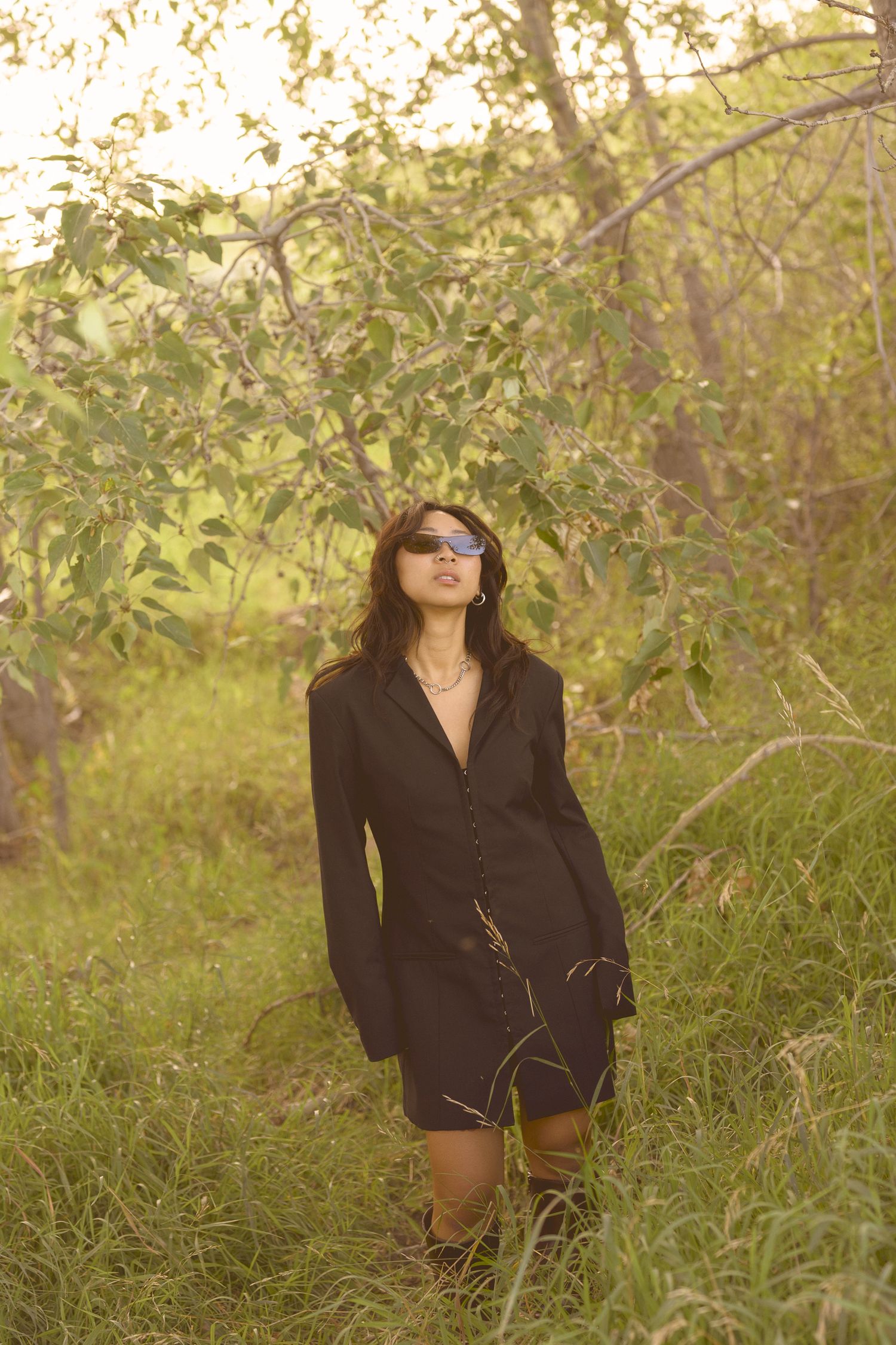 Fashion portrait of someone in black blazer dress posing among trees at golden hour.