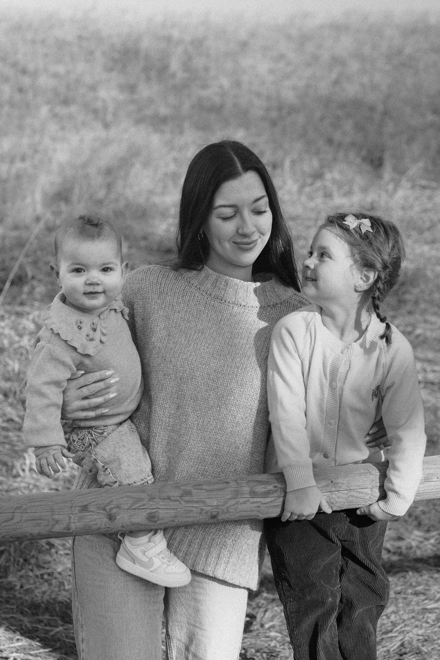 Black and white portrait of a family sharing a tender moment together outdoors while leaning against a wooden fence.