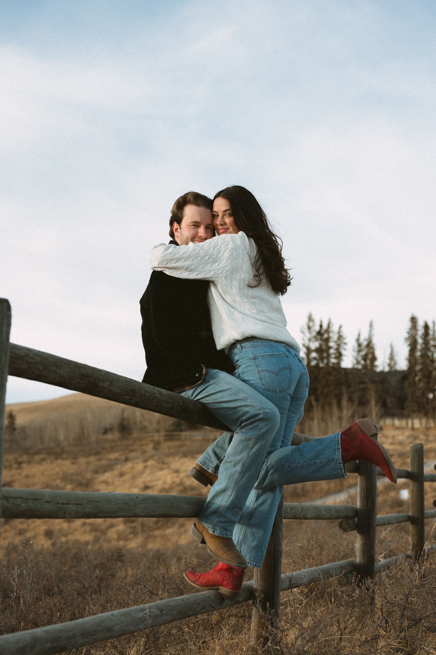 A tender moment shared between two people on a wooden fence with mountains and pine trees in the background.
