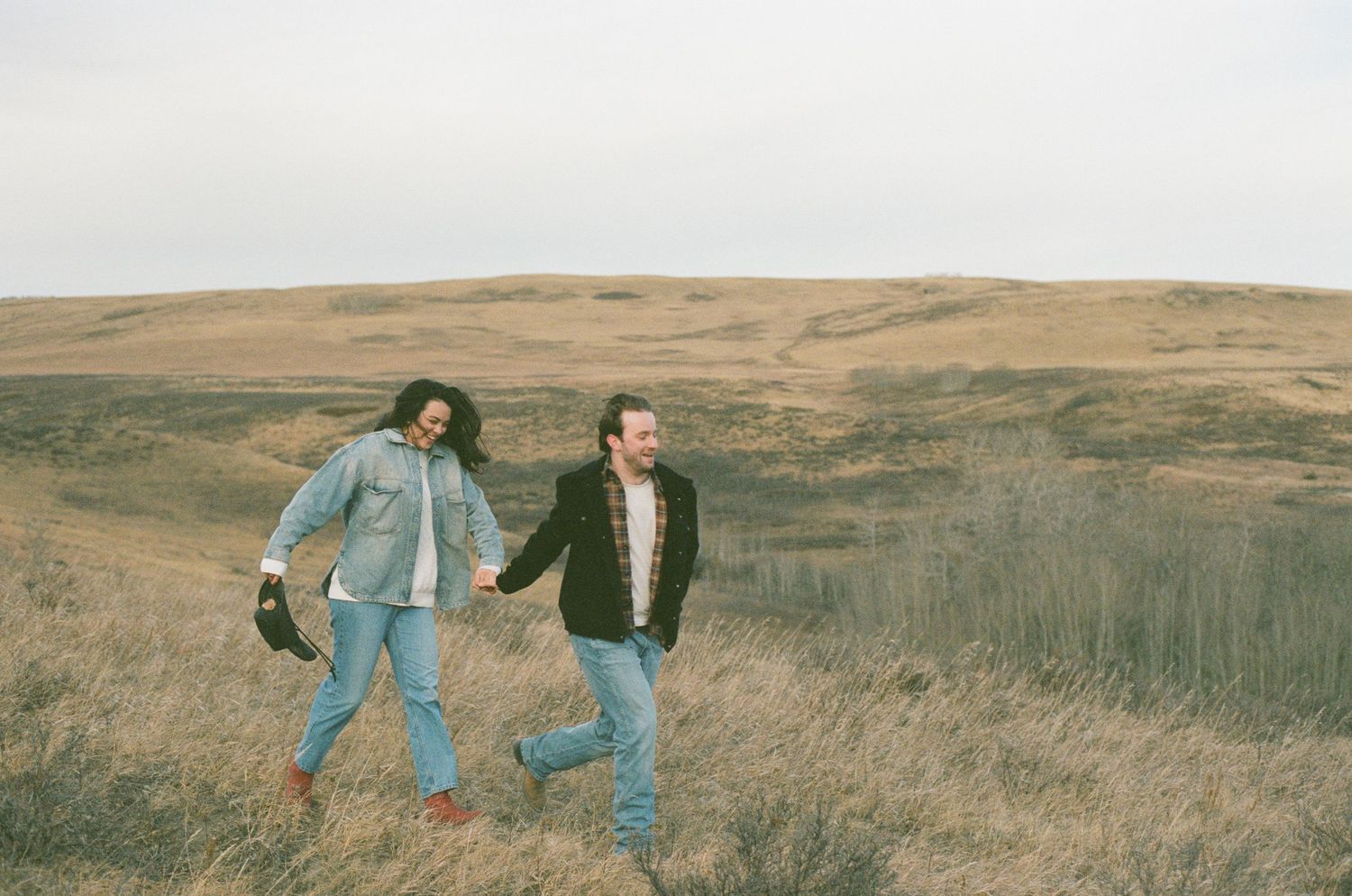 Two people in denim walk through a scenic autumn field with rolling hills in the background.