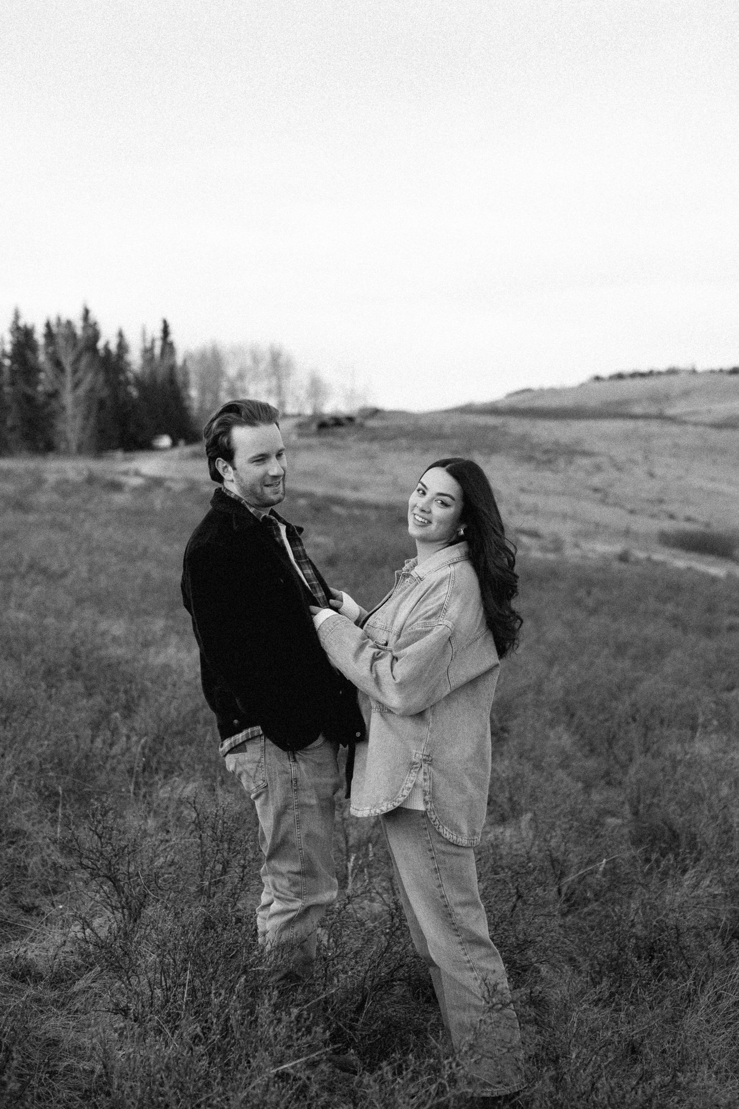 A couple shares a candid moment together in a black and white photo on a grassy hillside with trees in the background.