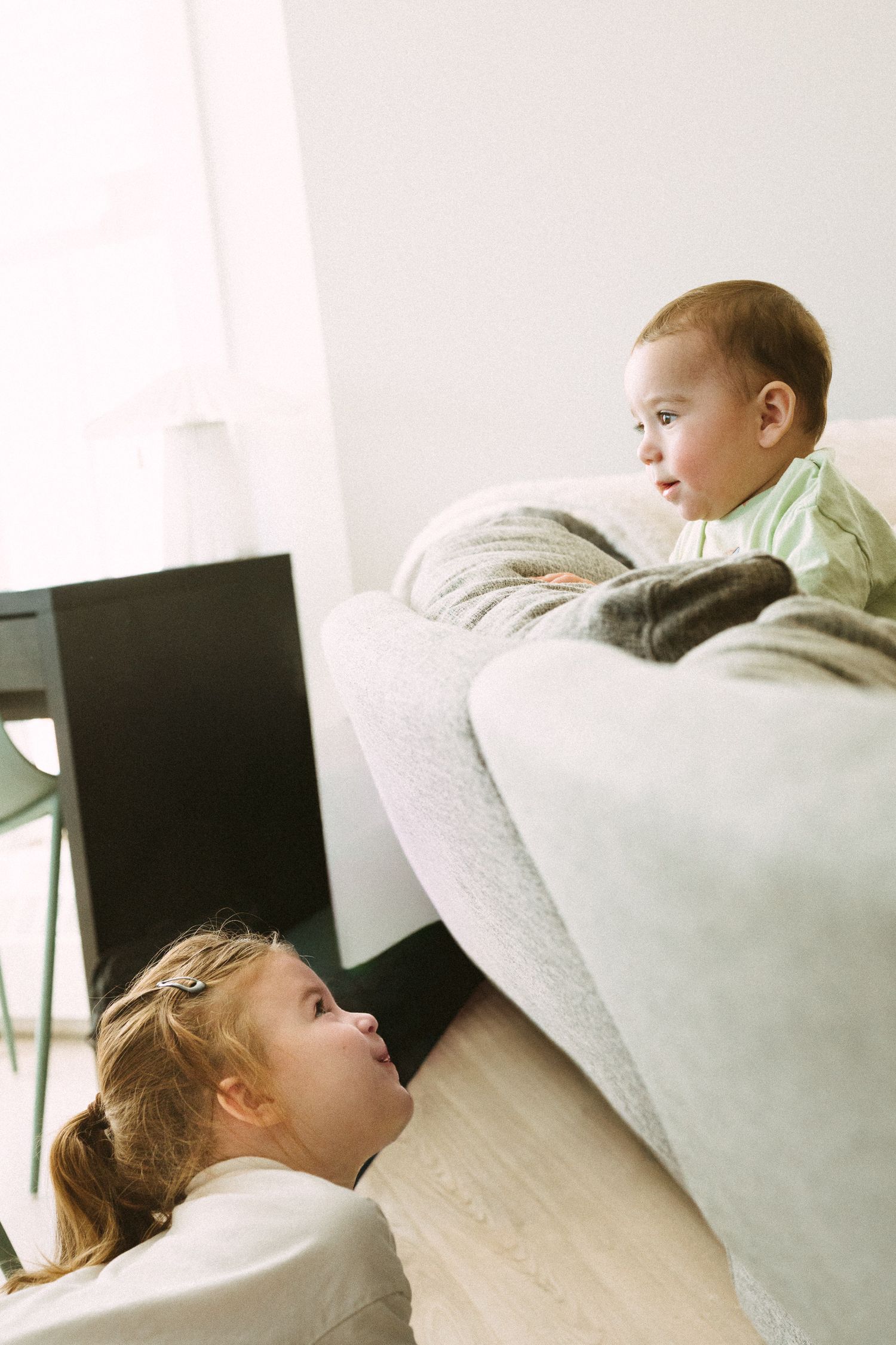 Two children share a playful moment near a couch, with one lying on the floor and another perched above.