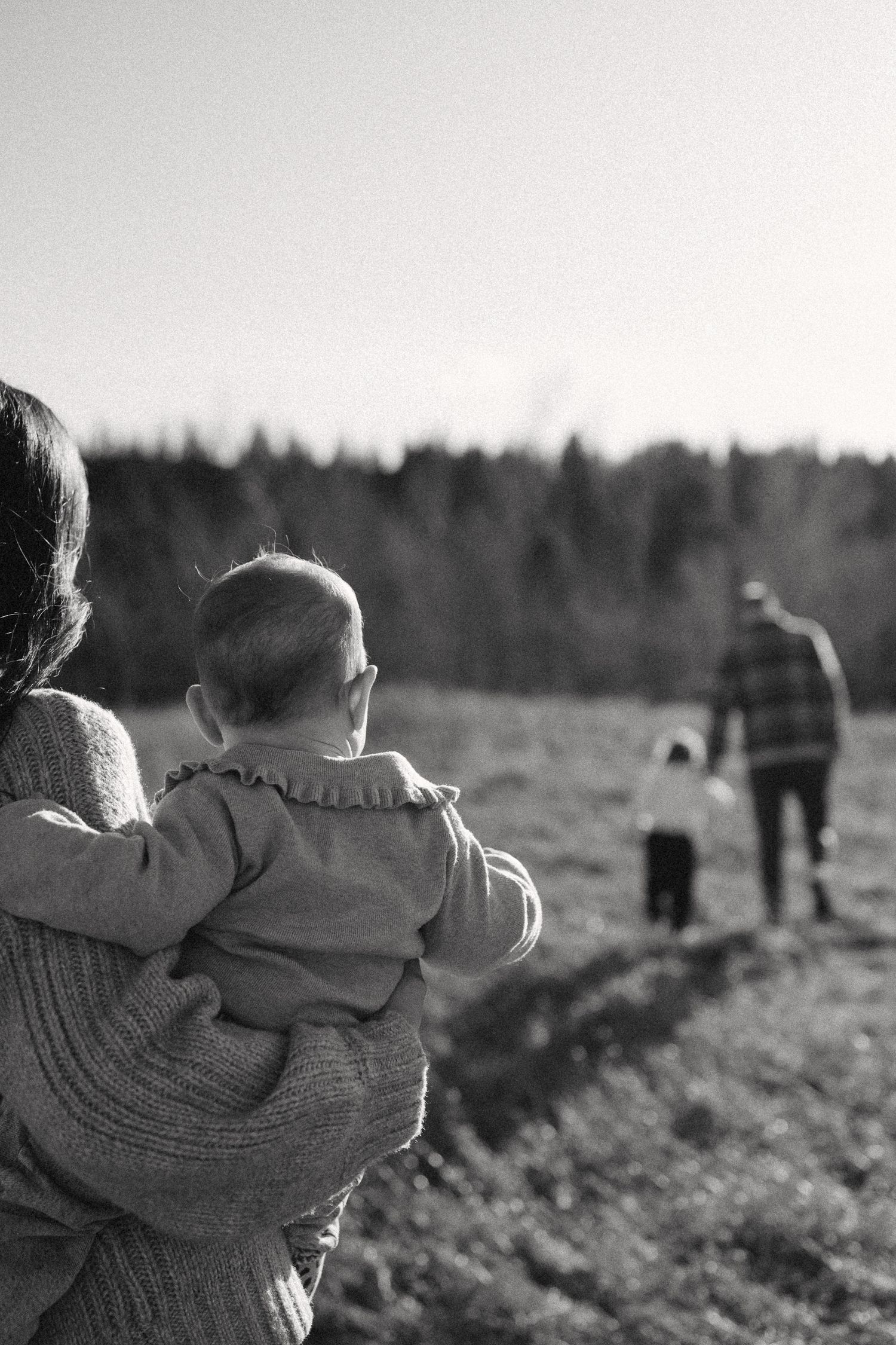 A monochrome image showing figures walking through a rural field with forest in the distance.