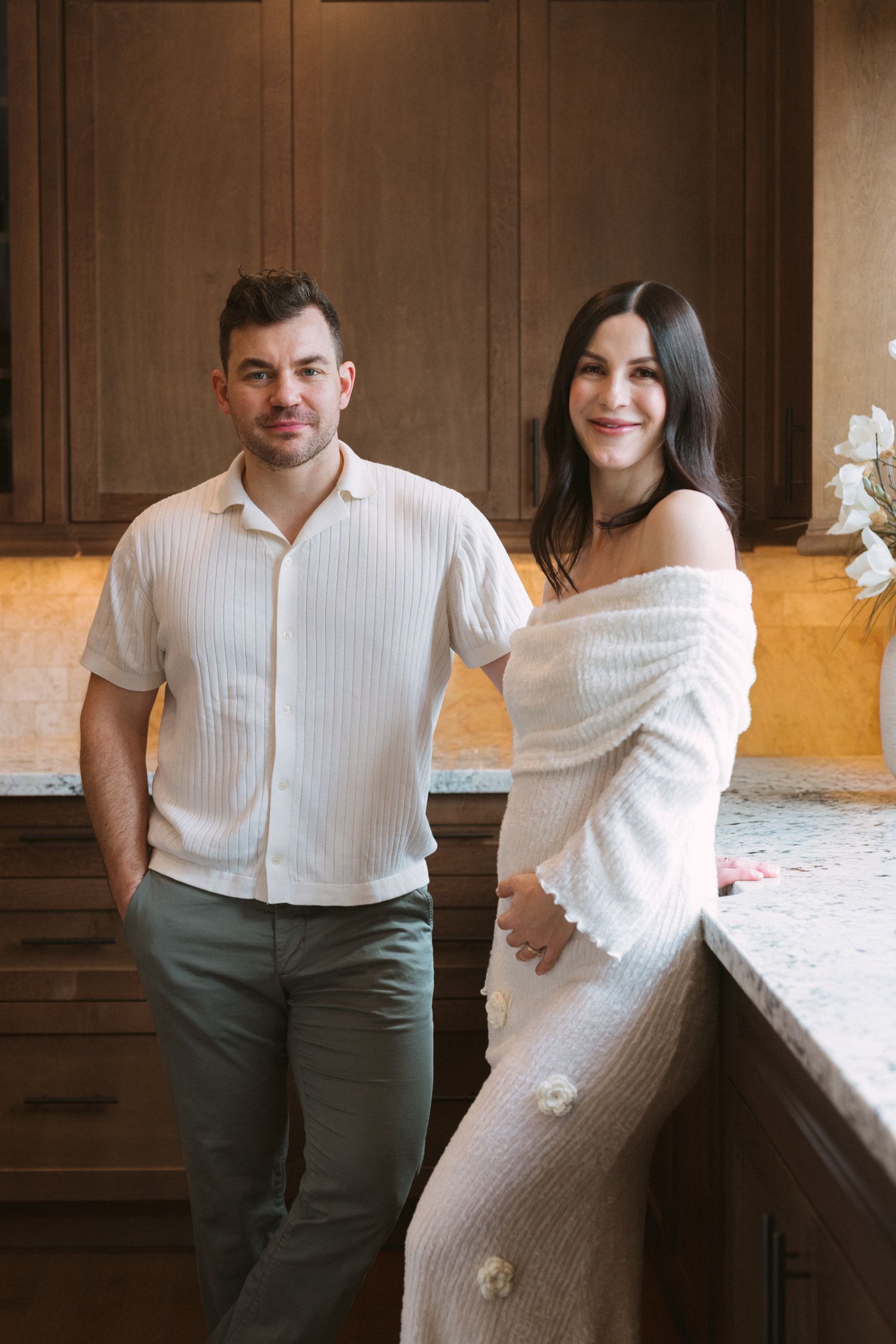 A couple smiles together in a warm kitchen, woman in off-shoulder white dress, man in cream shirt and sage pants.