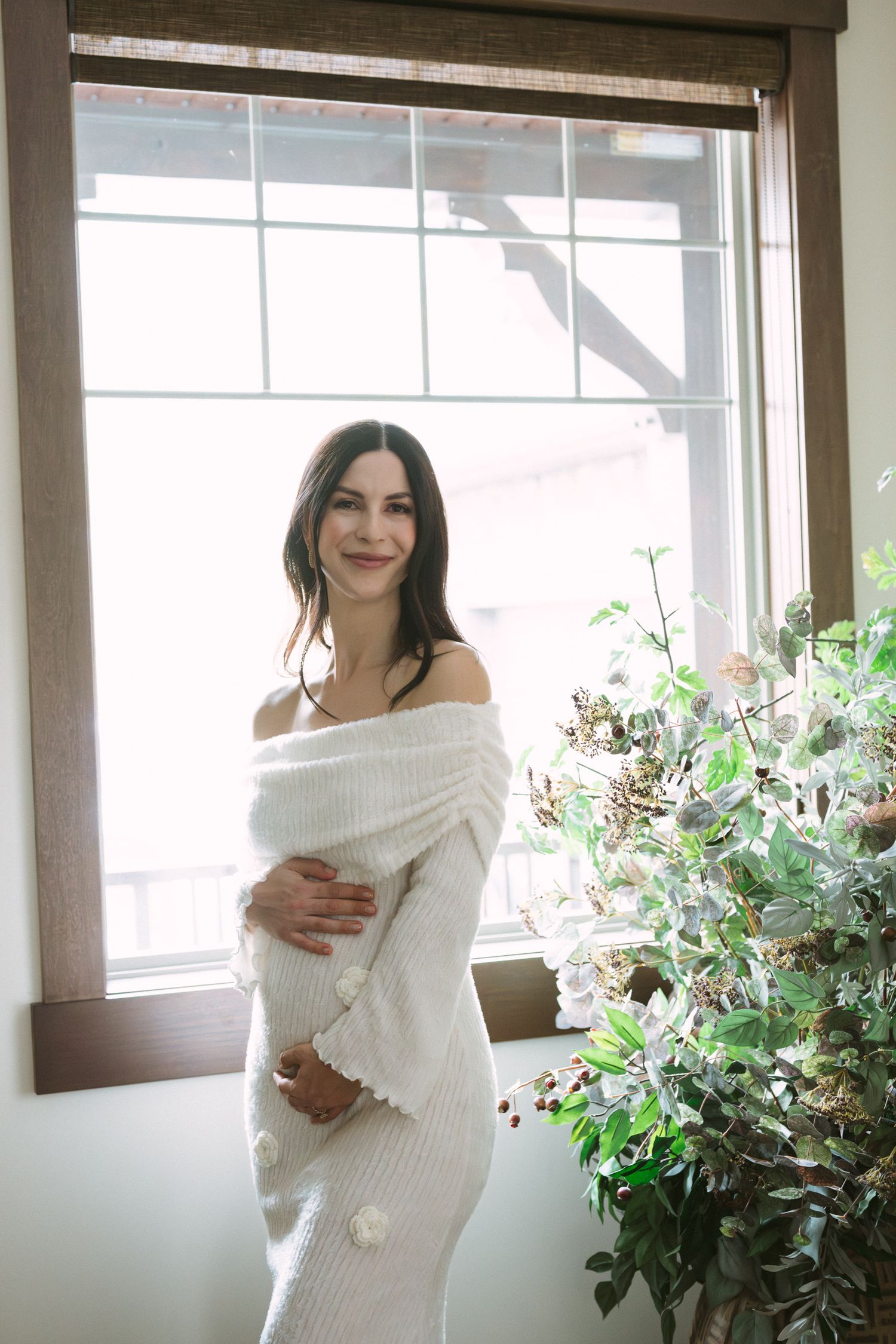 Pregnant woman in white off-shoulder knit dress smiling by a window with greenery nearby.