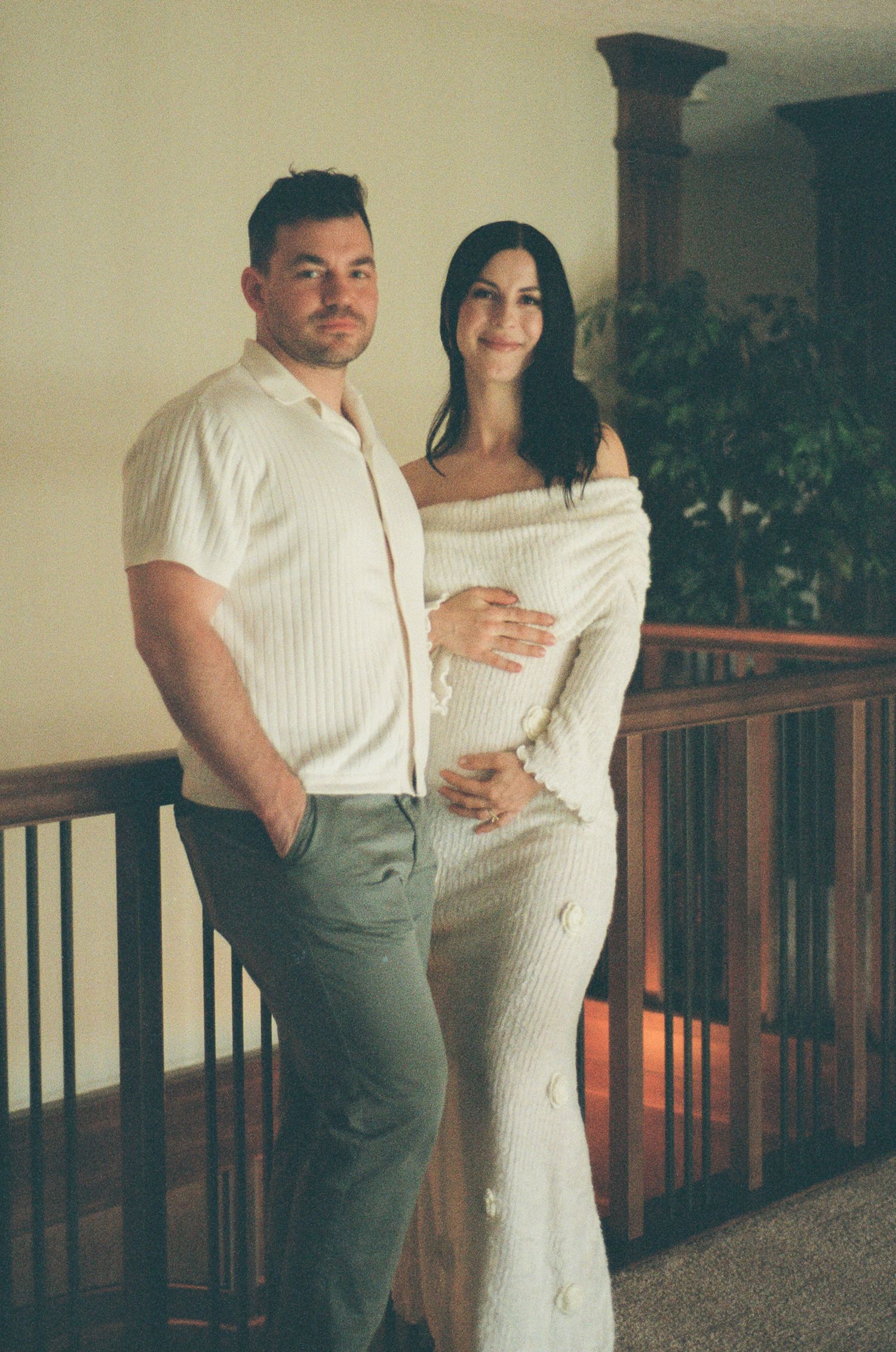 Couple stands by wooden railing, pregnant woman in white knit dress beside man in cream polo.