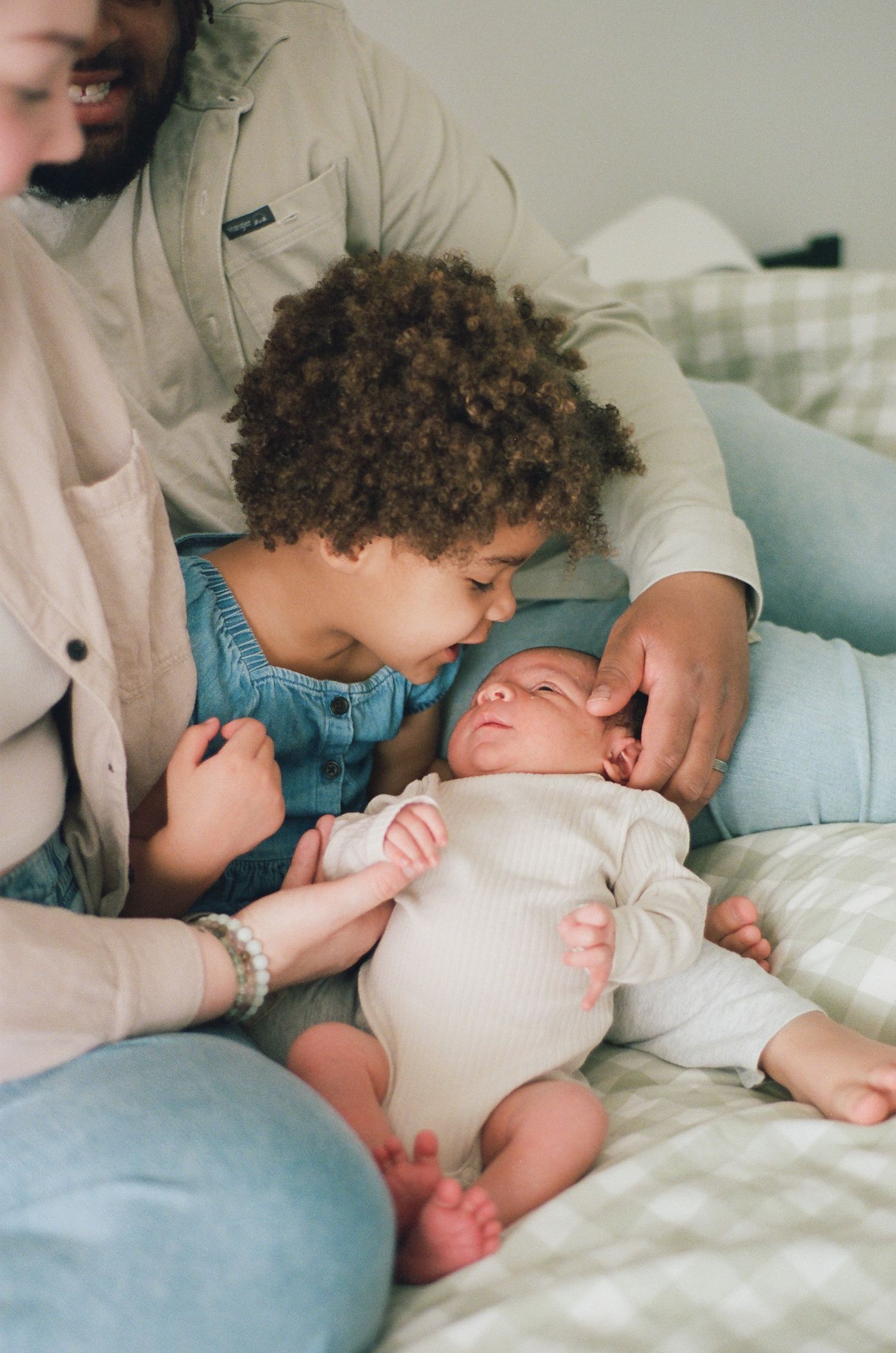 Older toddler sibling gently kissing newborn baby lying on bed surrounded by loving family.