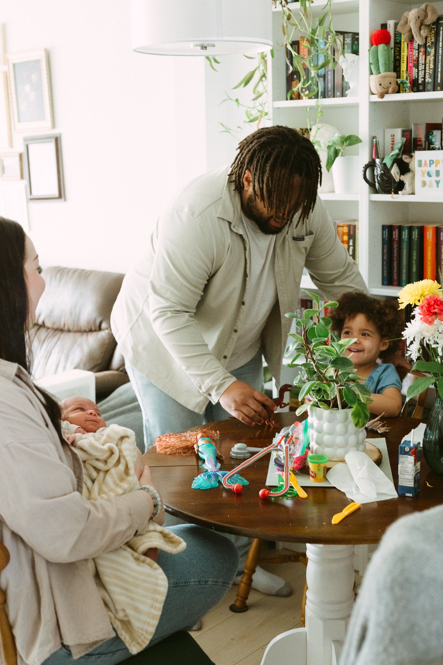 Father playing with toddler son at dining table while mother holds newborn baby in cozy home.