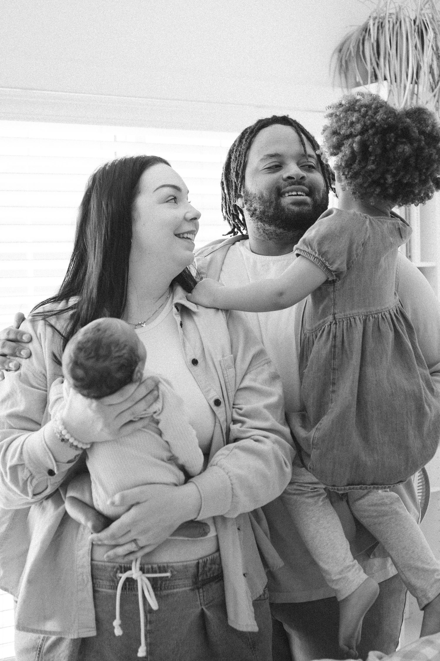 Black and white photo of a happy interracial family with father holding toddler daughter and mother cradling newborn baby.