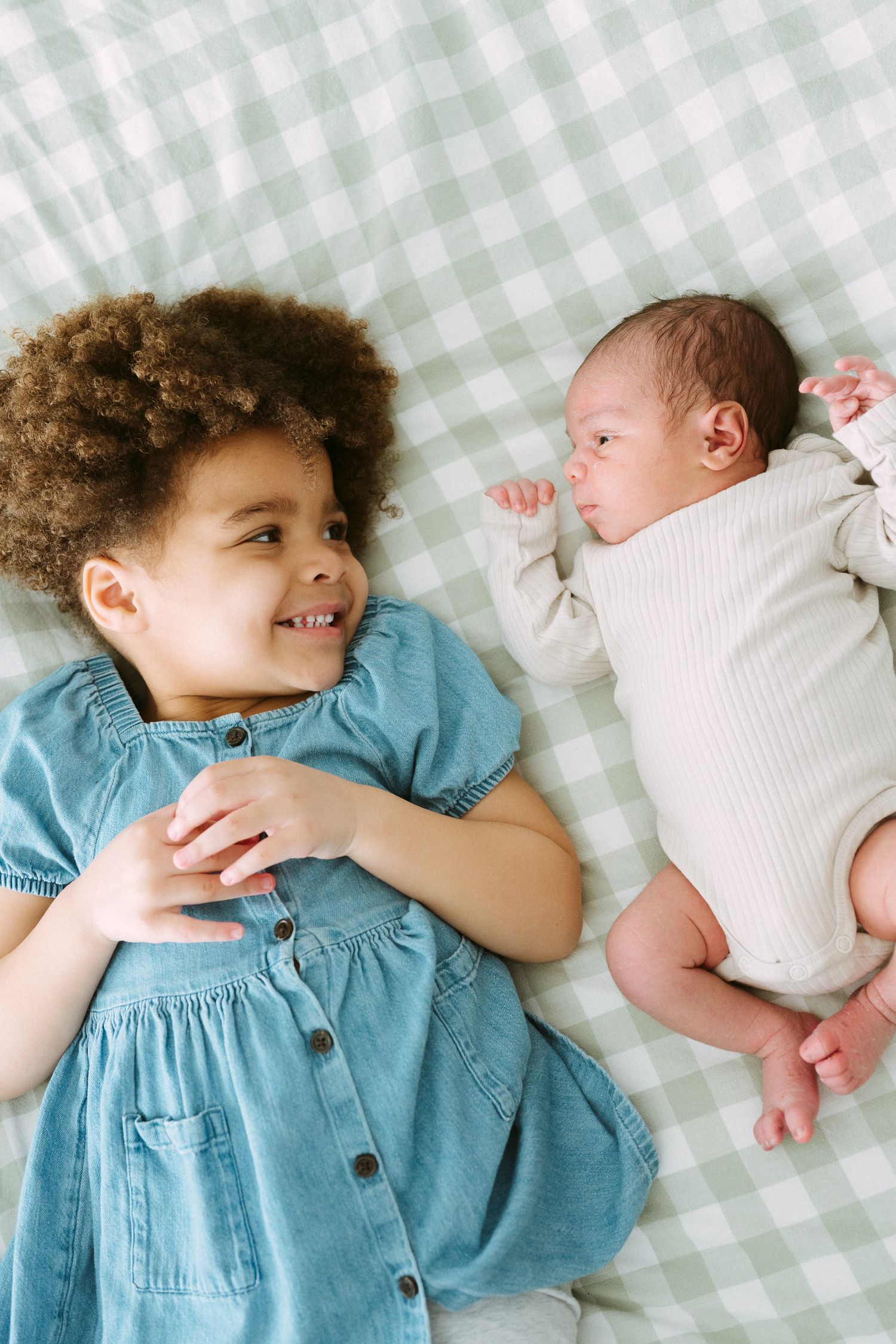Smiling curly-haired toddler in denim dress lies beside newborn baby on green gingham blanket.