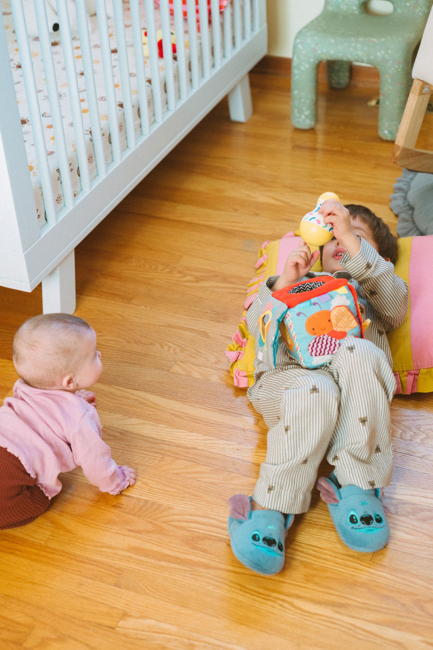 Children playing on hardwood floor near white crib with toys scattered around.