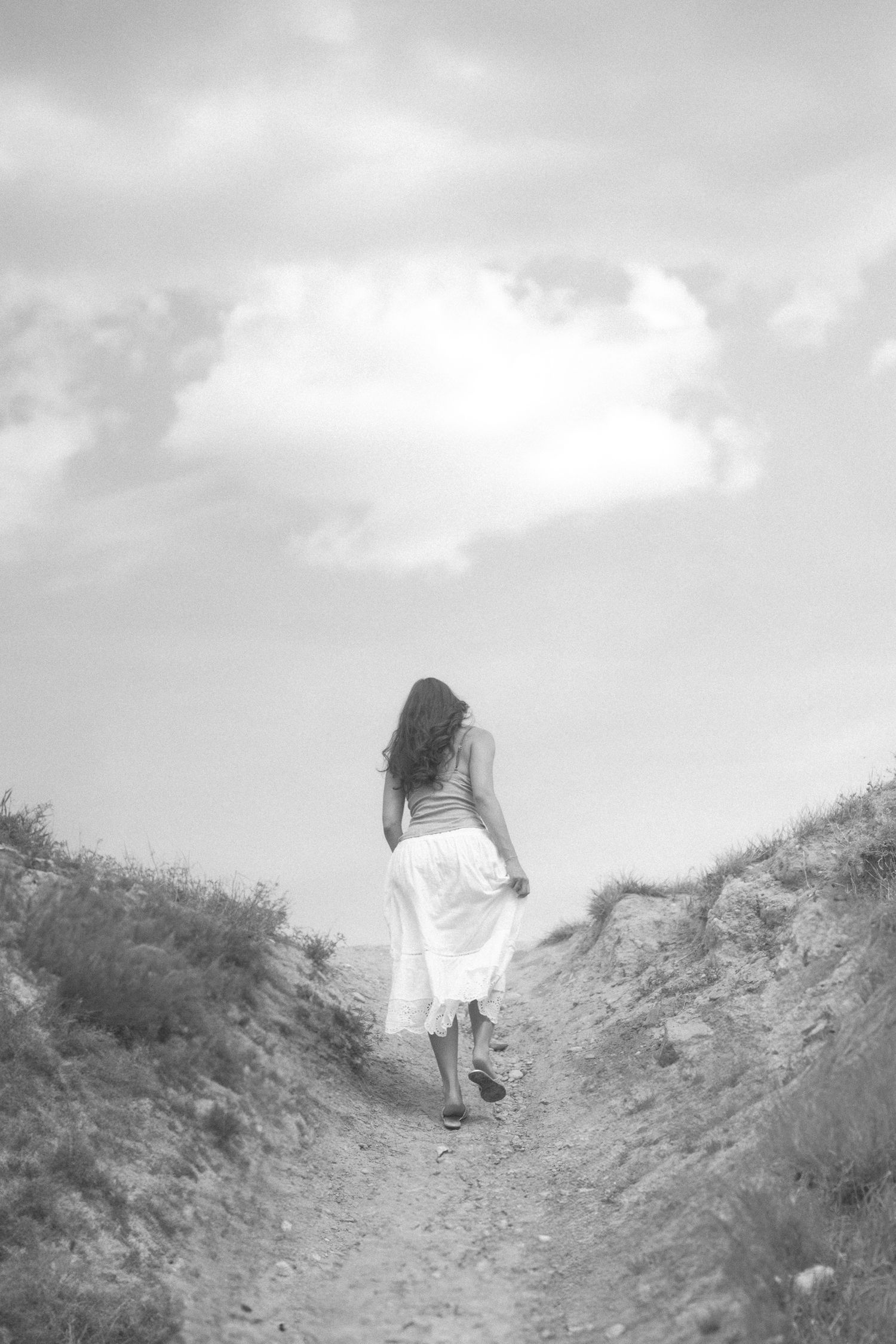 A figure in a white dress walks along a winding dirt path through grassy hills.
