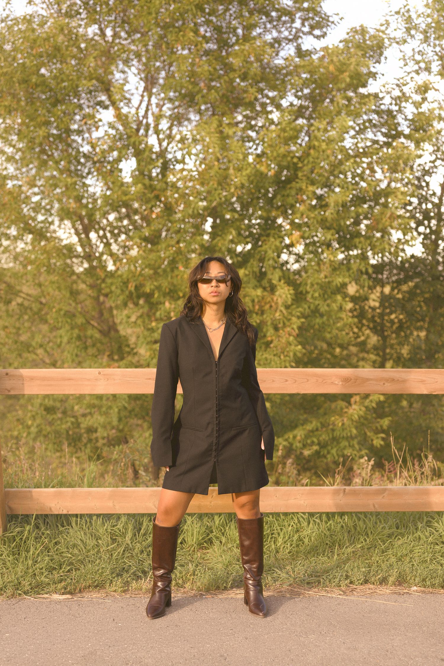 A figure in a black blazer and boots poses on a wooden bench in an outdoor setting with autumn trees.