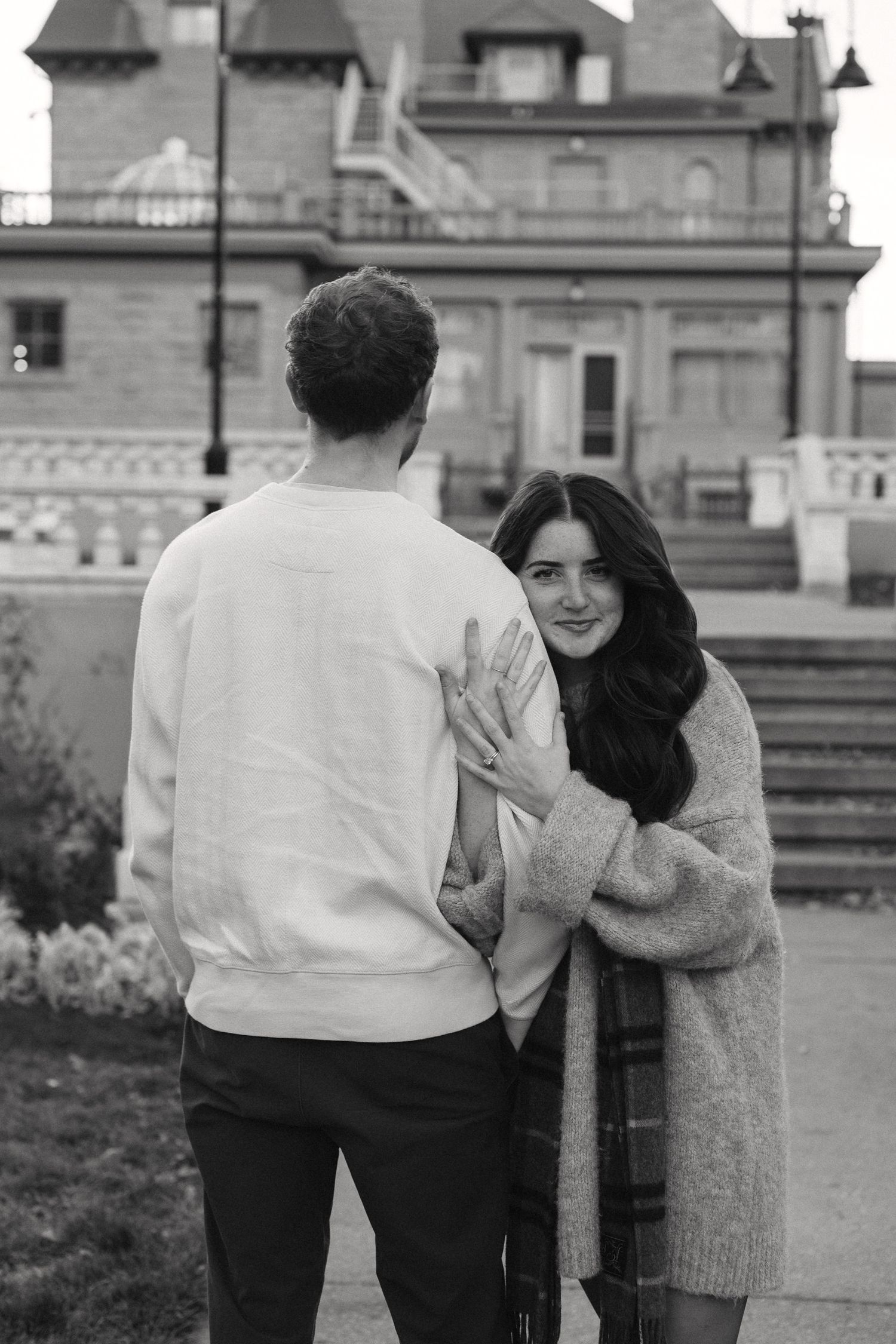 Black and white photo of a couple sharing a sweet moment outdoors with urban architecture behind them.