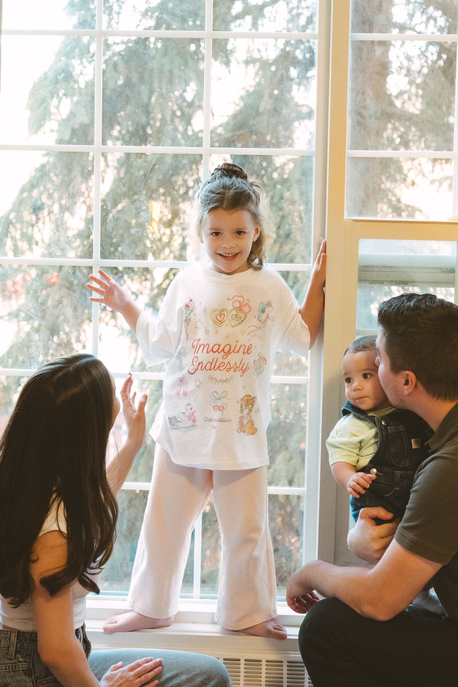 Family gathering by large window with natural light streaming in as young child stands on windowsill in casual attire.