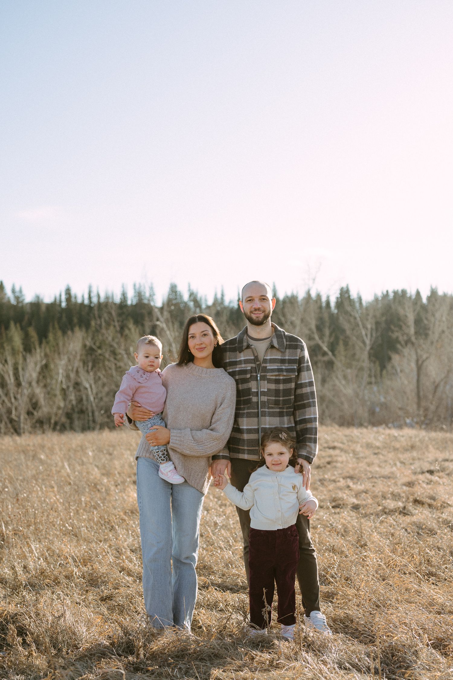 A family poses together in a golden field during sunset with evergreen trees in the background.