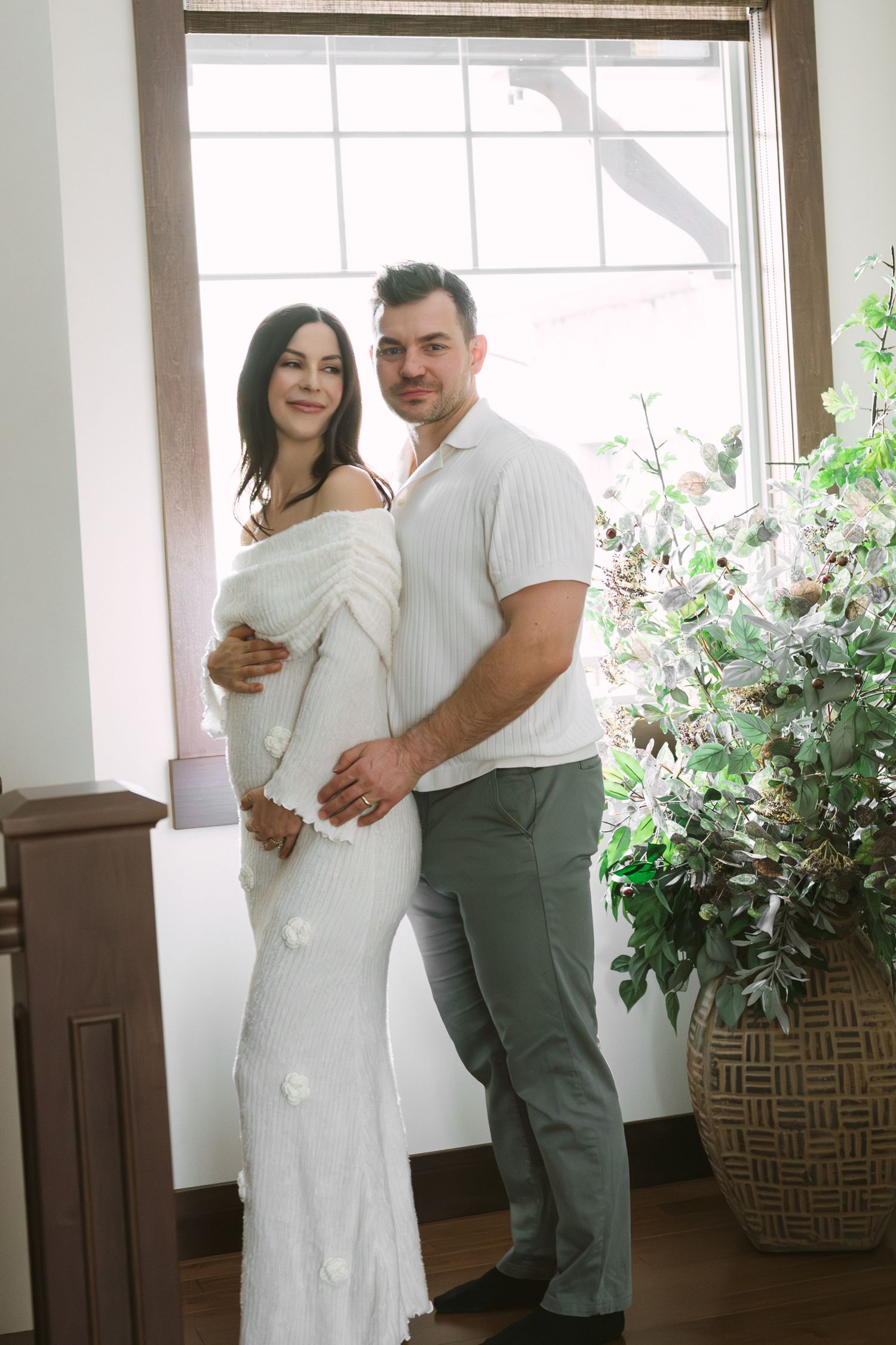 Couple poses together indoors, pregnant woman in white off-shoulder dress embraced by partner near window.