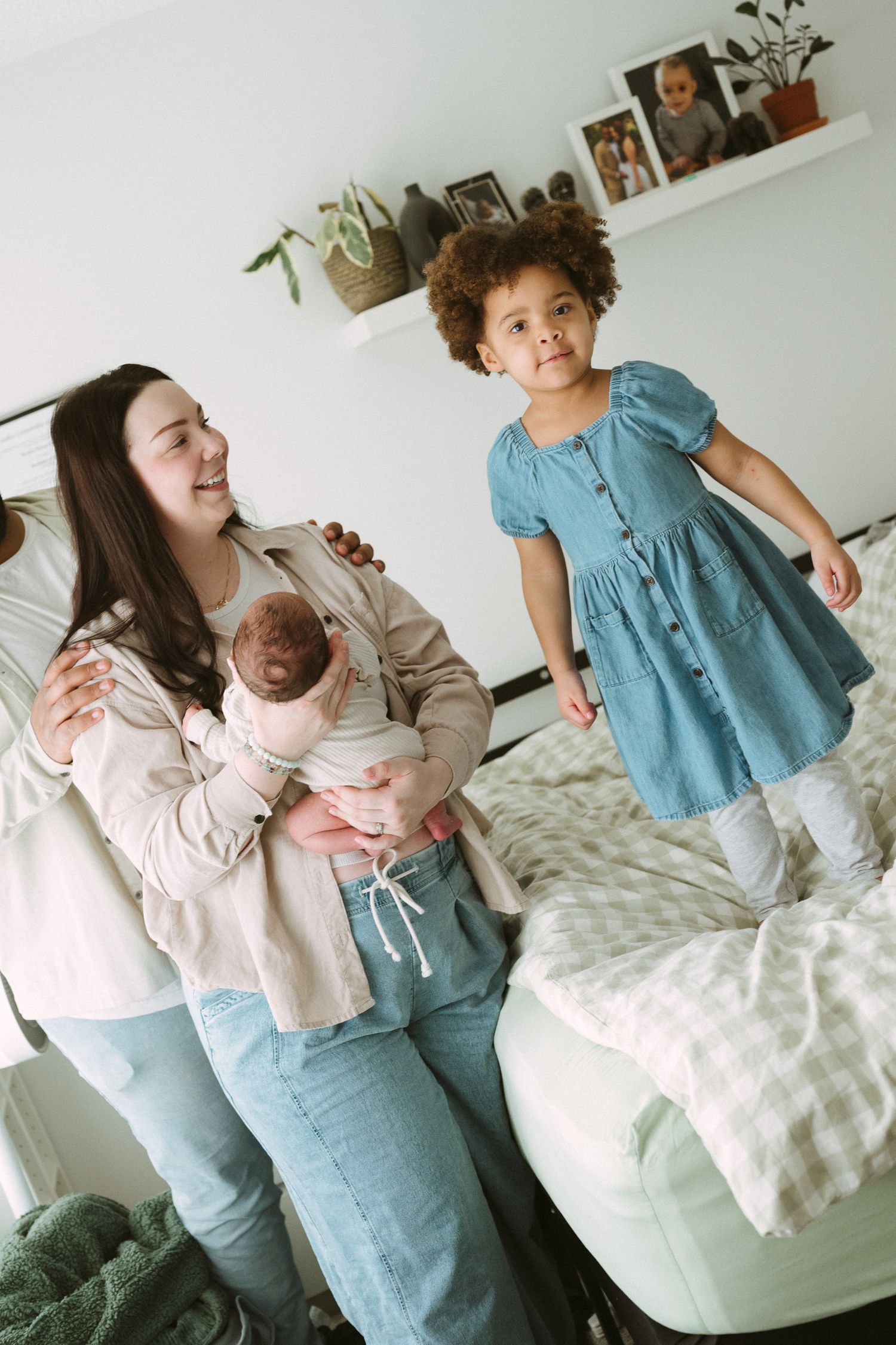 Multiracial family with two mothers holding a newborn baby while their young daughter stands on a bed in a bright bedroom.