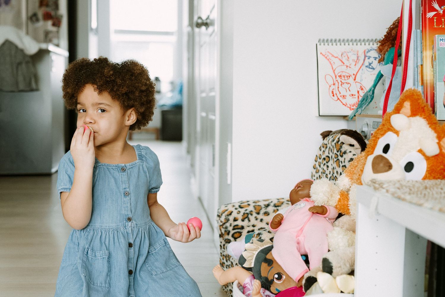 A young girl in a blue dress stands near a shelf of stuffed animals, covering her mouth with her hand.