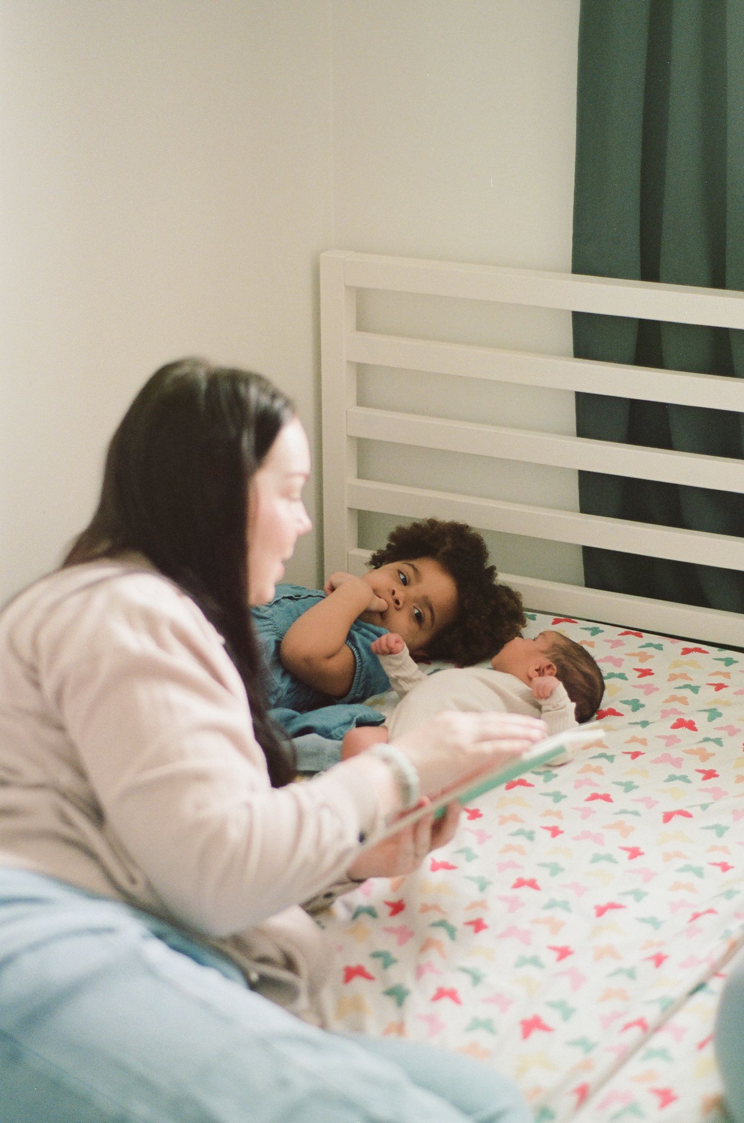A mother reads to her toddler and newborn baby lying on a colorful patterned bed in a cozy bedroom.