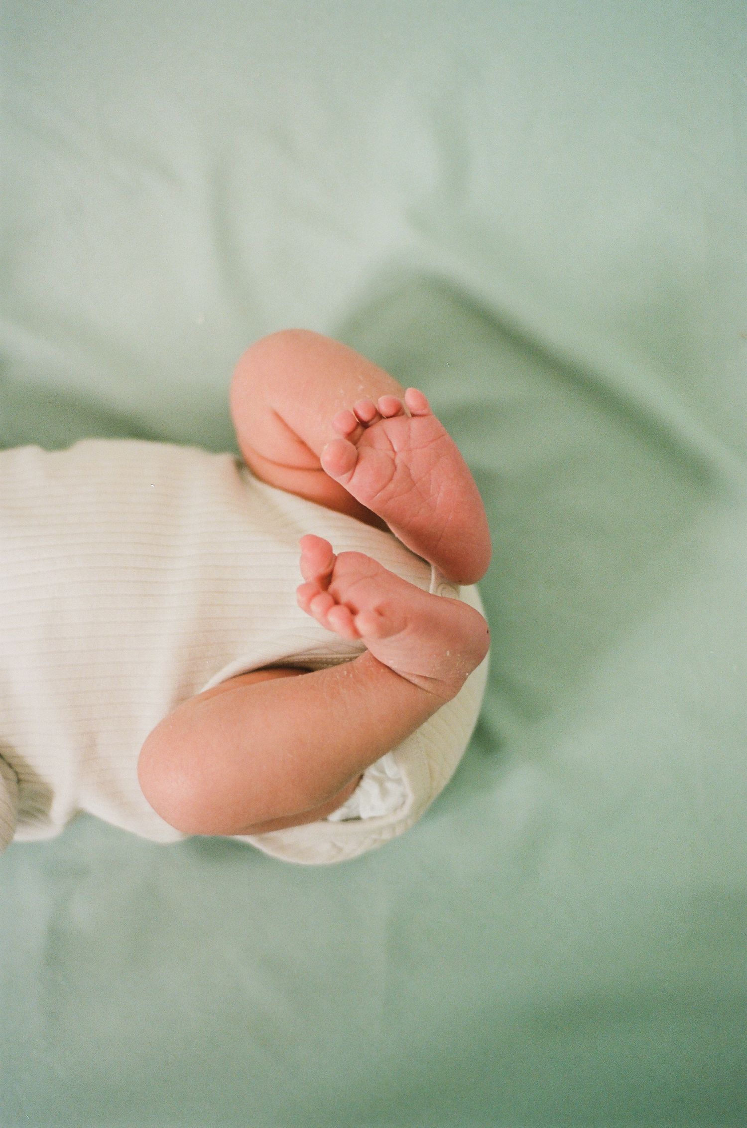 Close-up of newborn baby feet and legs in white ribbed onesie on soft green blanket.