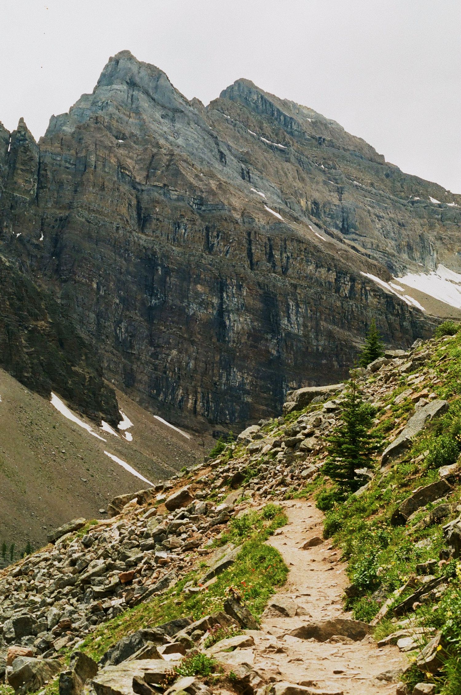 A hiking trail traverses rocky terrain beneath massive mountain cliffs with patches of snow.
