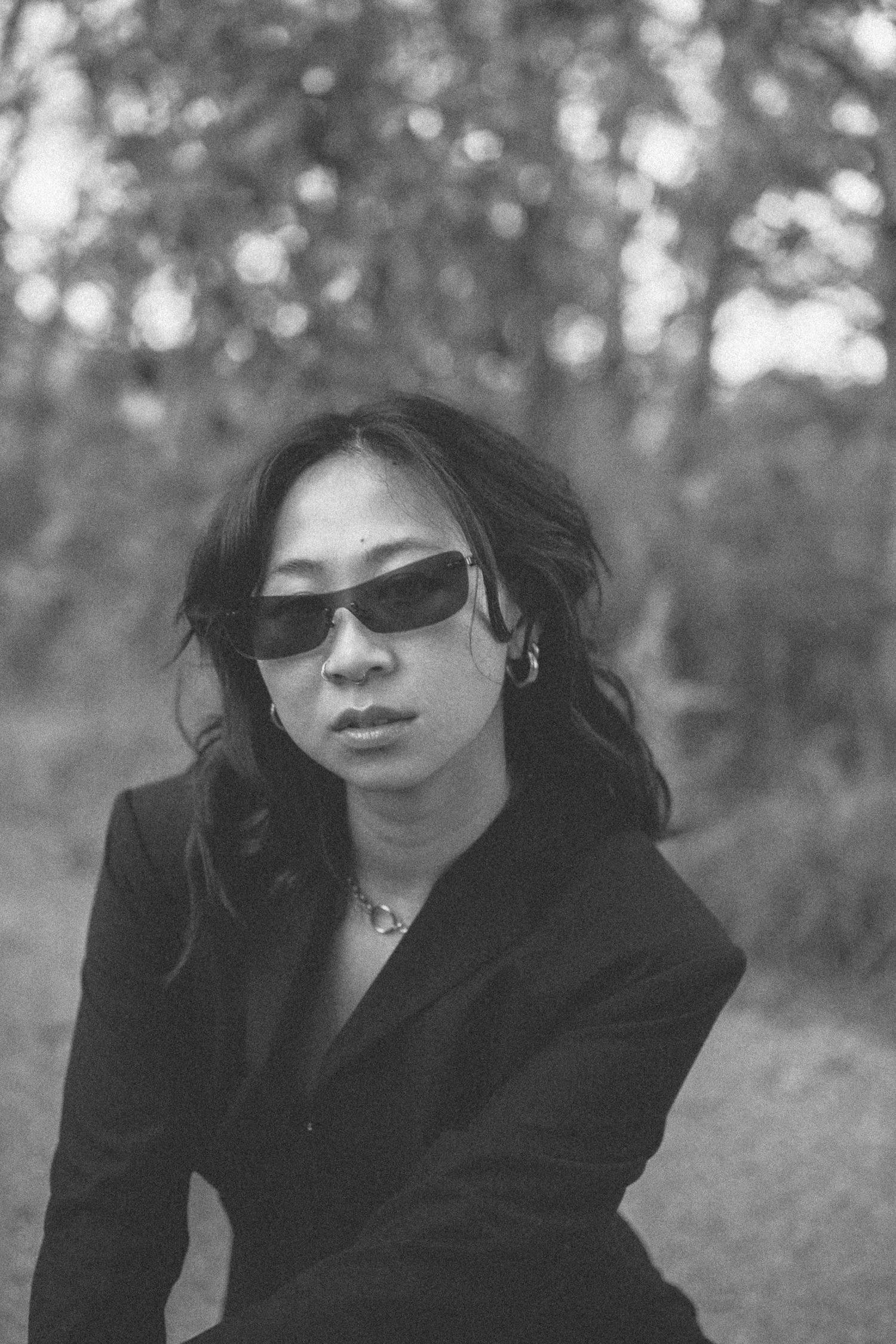 Black and white portrait of person in sunglasses and blazer with natural bokeh background.