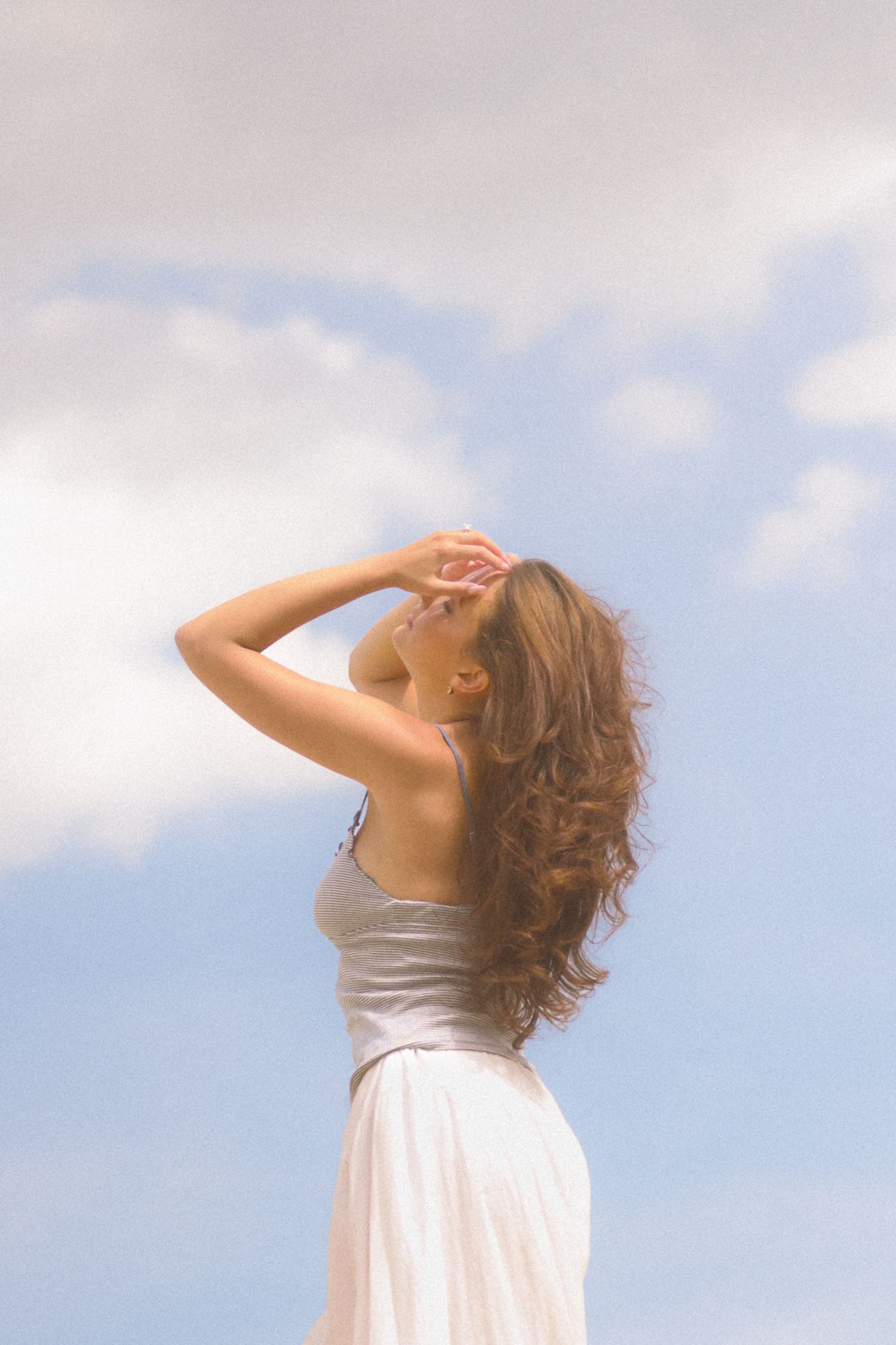 A person in a light dress tilts their head back against a cloudy blue sky background.