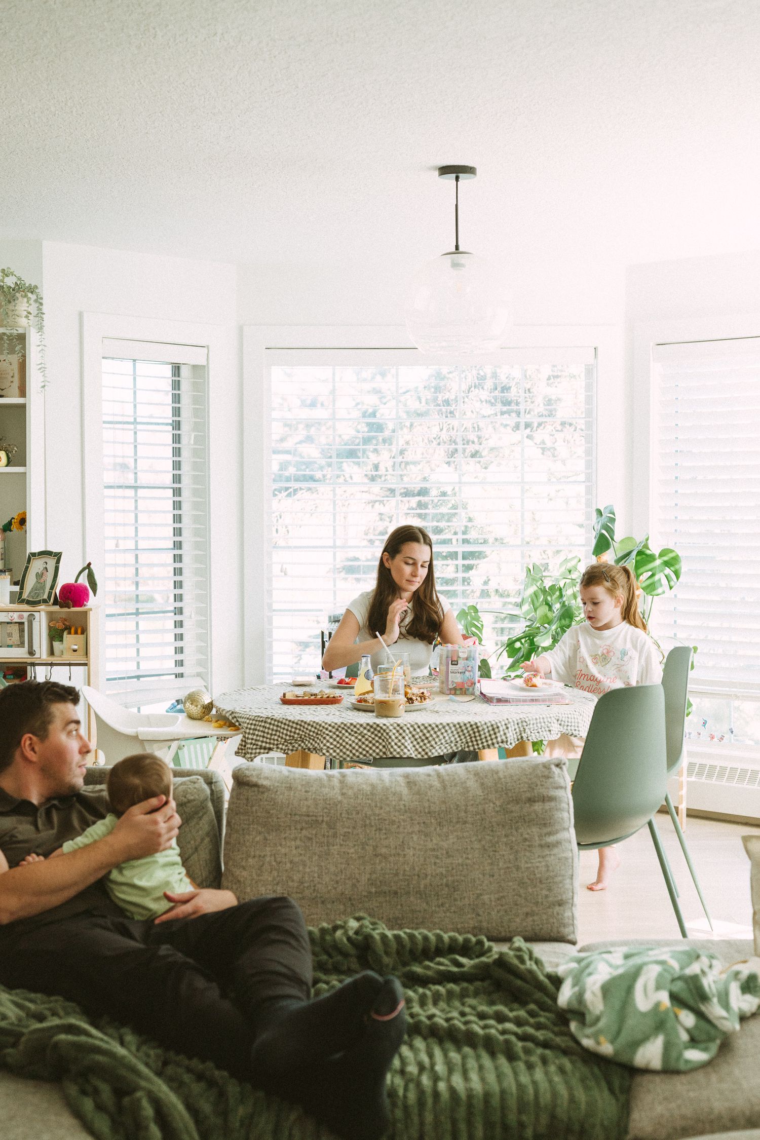 A bright living room scene with a family gathered around a dining table and relaxing on a couch by large windows.