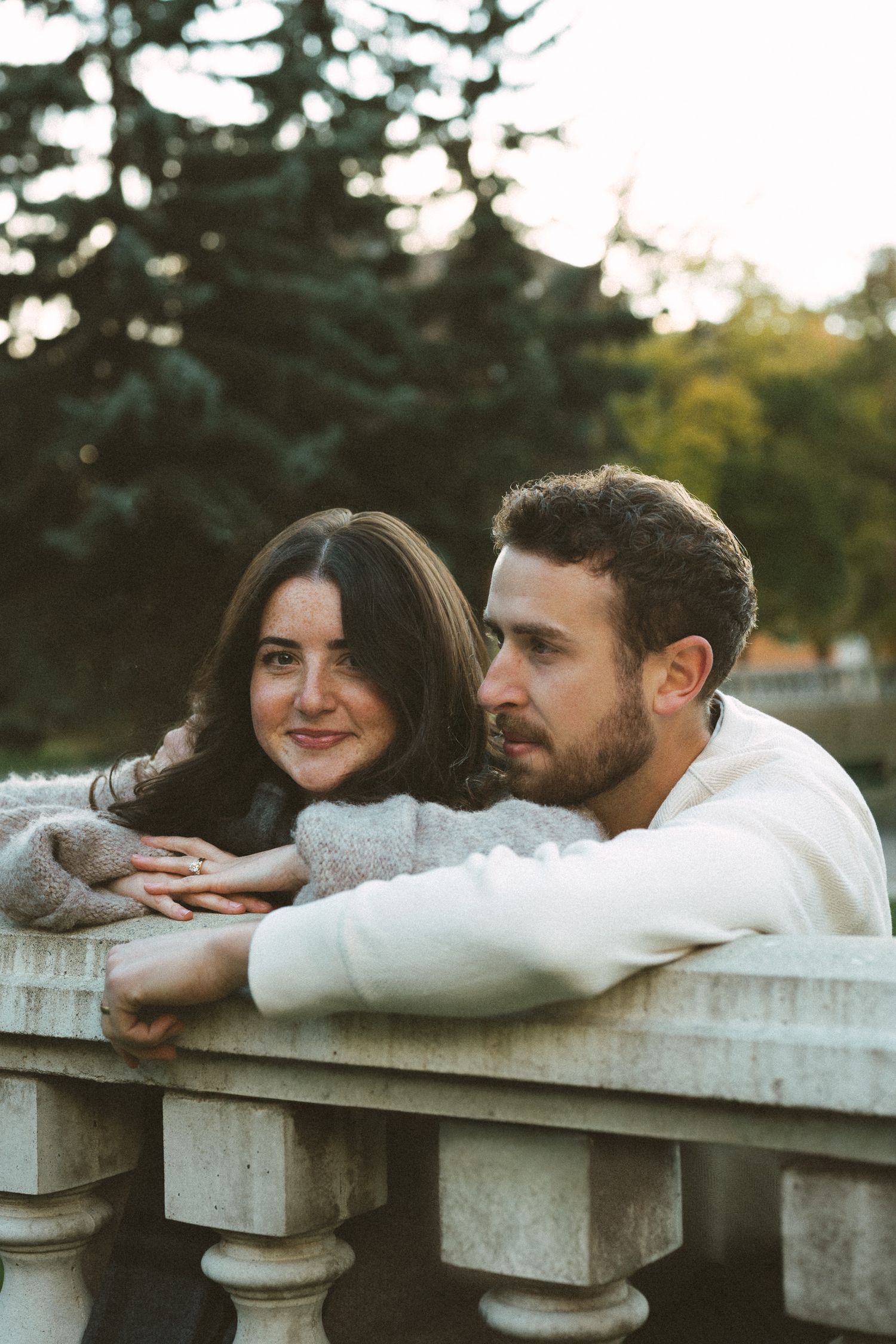 Couple leaning on a stone balustrade together during sunset with evergreen trees in background.