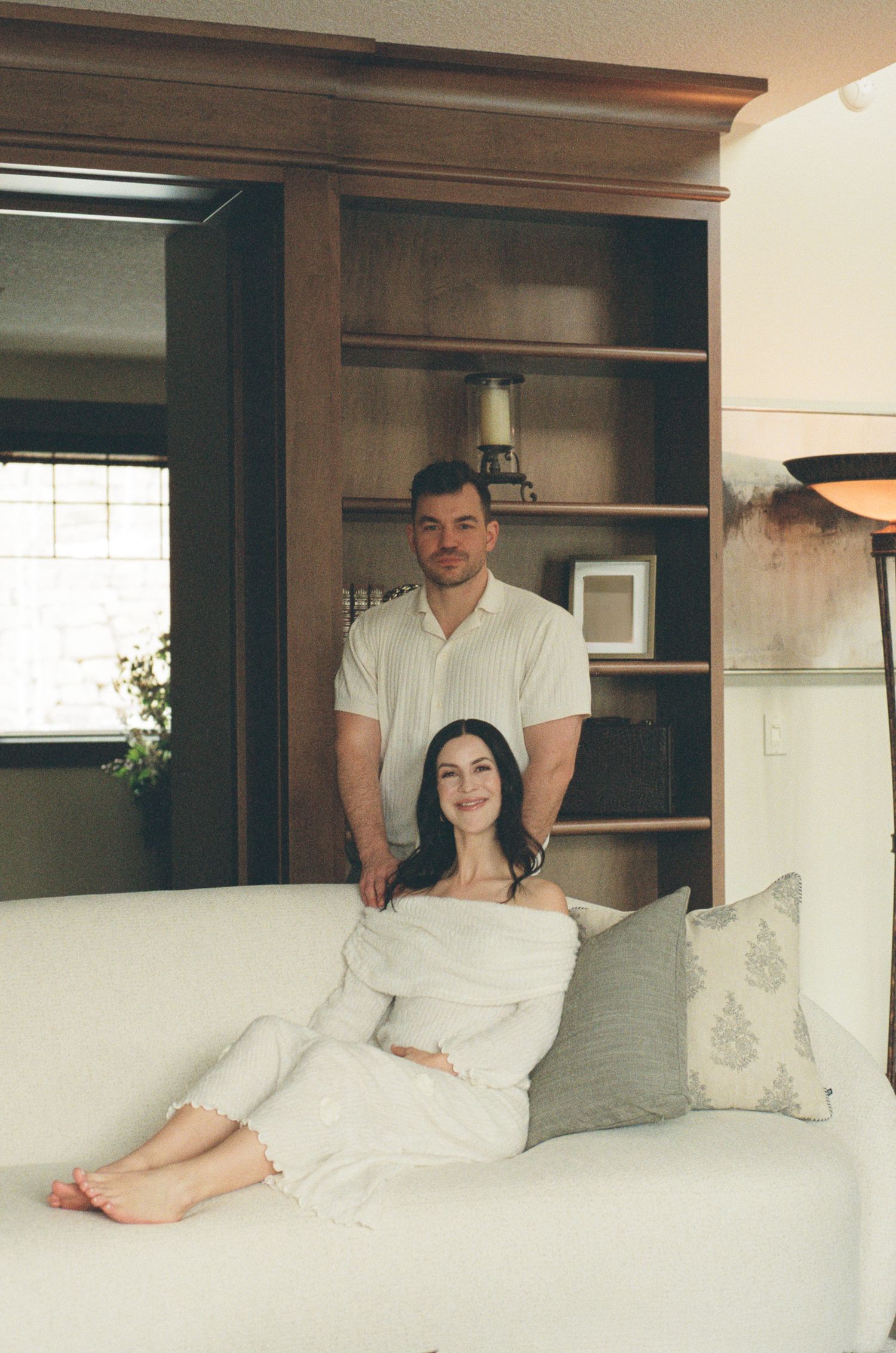 A couple poses in a cozy living room, woman sitting on white sofa, man standing behind her near wooden bookshelf.