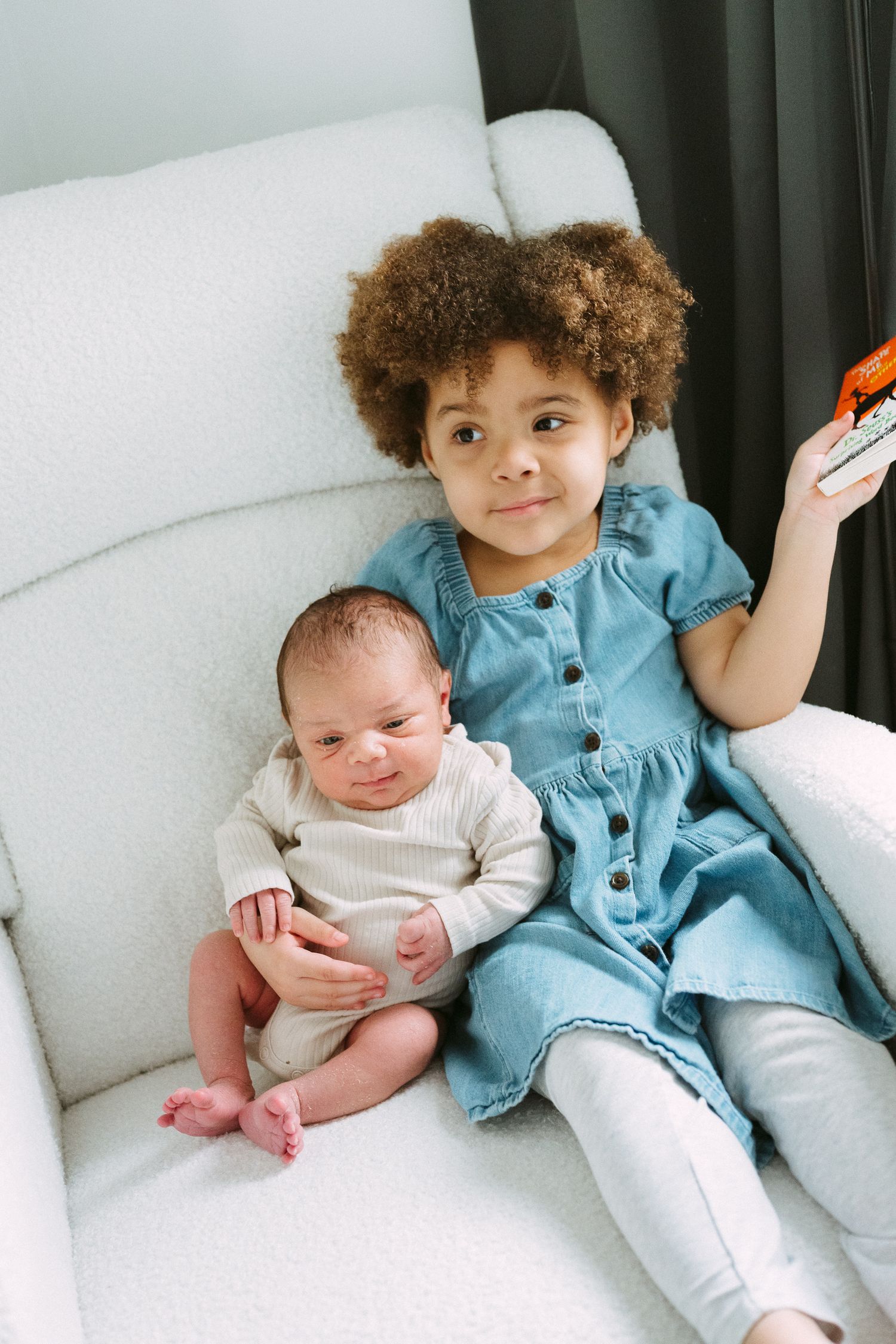 Smiling curly-haired toddler in blue denim dress sits on white chair holding newborn sibling and small toy.