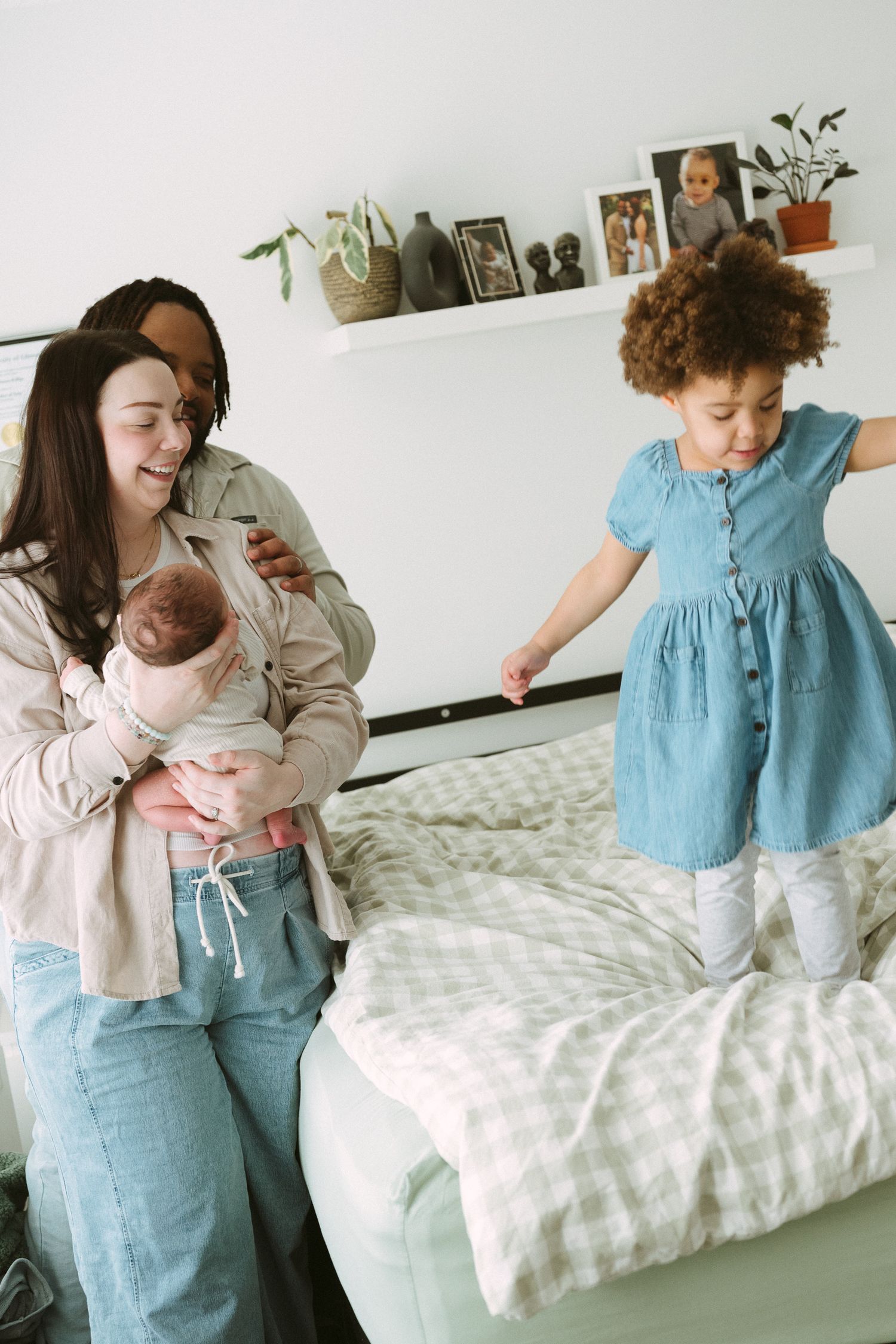Two mothers with a newborn baby and toddler girl in blue dress jumping on bed in bright bedroom.
