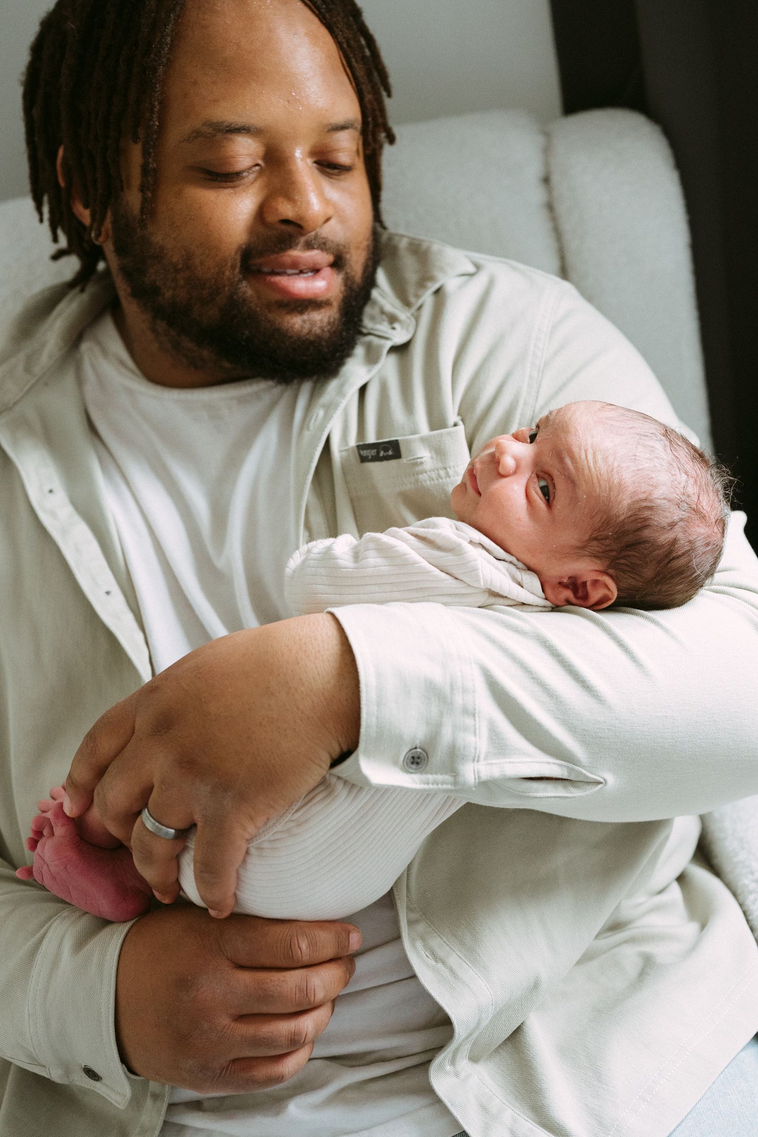 A father tenderly cradles his newborn baby in his arms, wearing a beige shirt and a wedding ring.