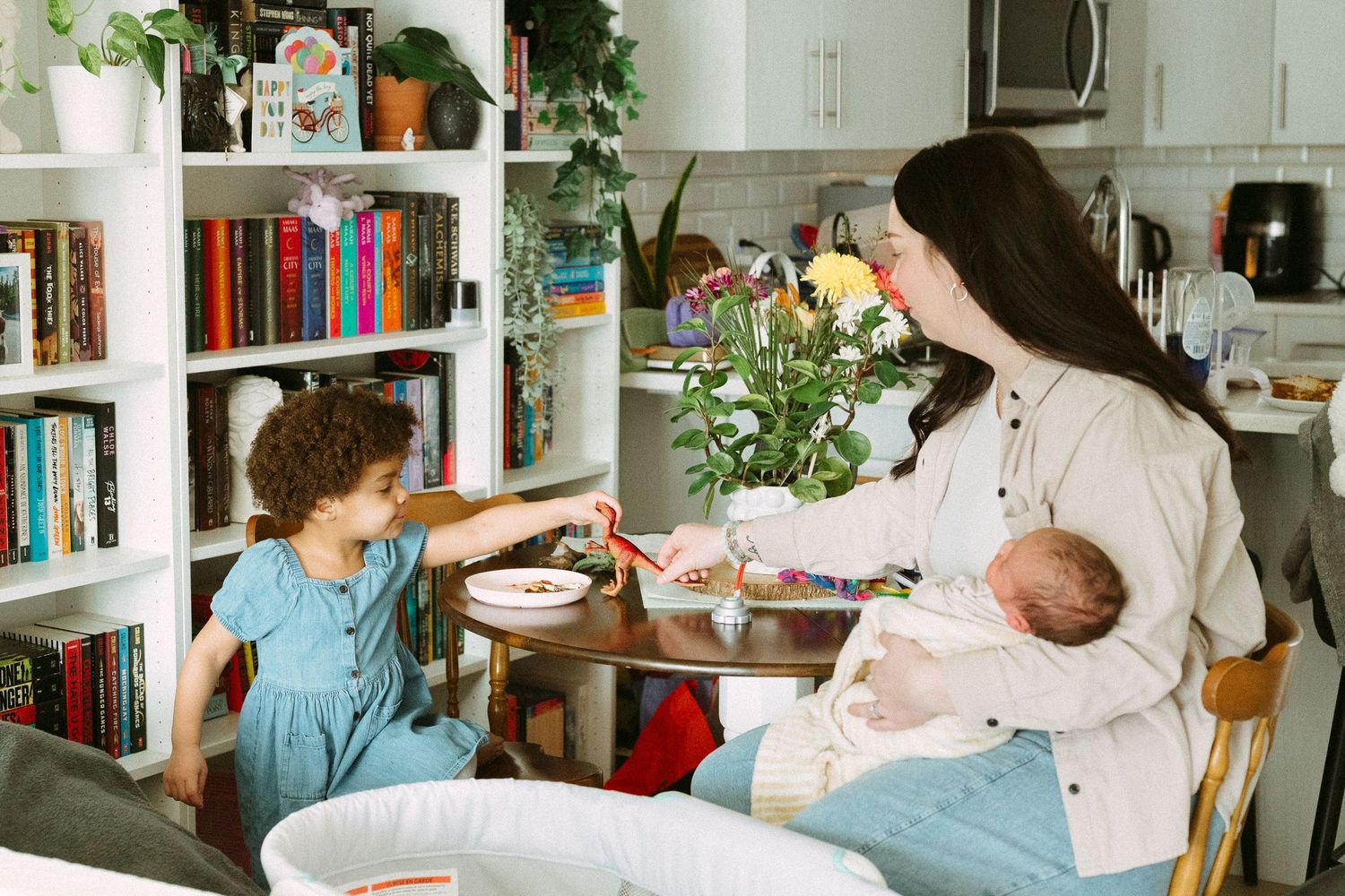 Mother holds newborn while interacting with young child at dining table in cozy home with bookshelf.