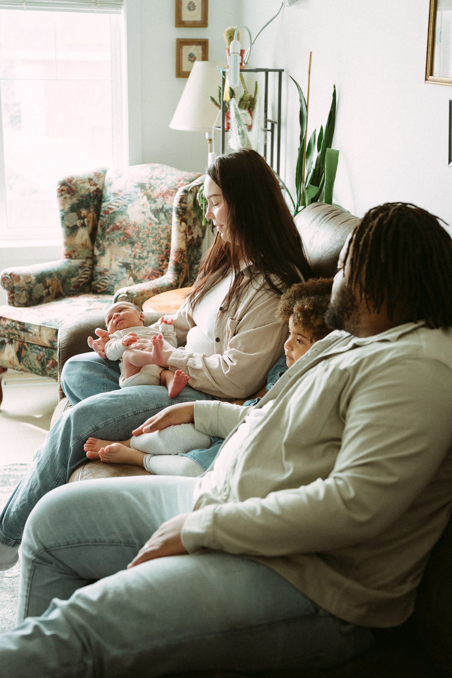 A young interracial family sits together on a couch, with a mother cradling a newborn baby beside her partner.
