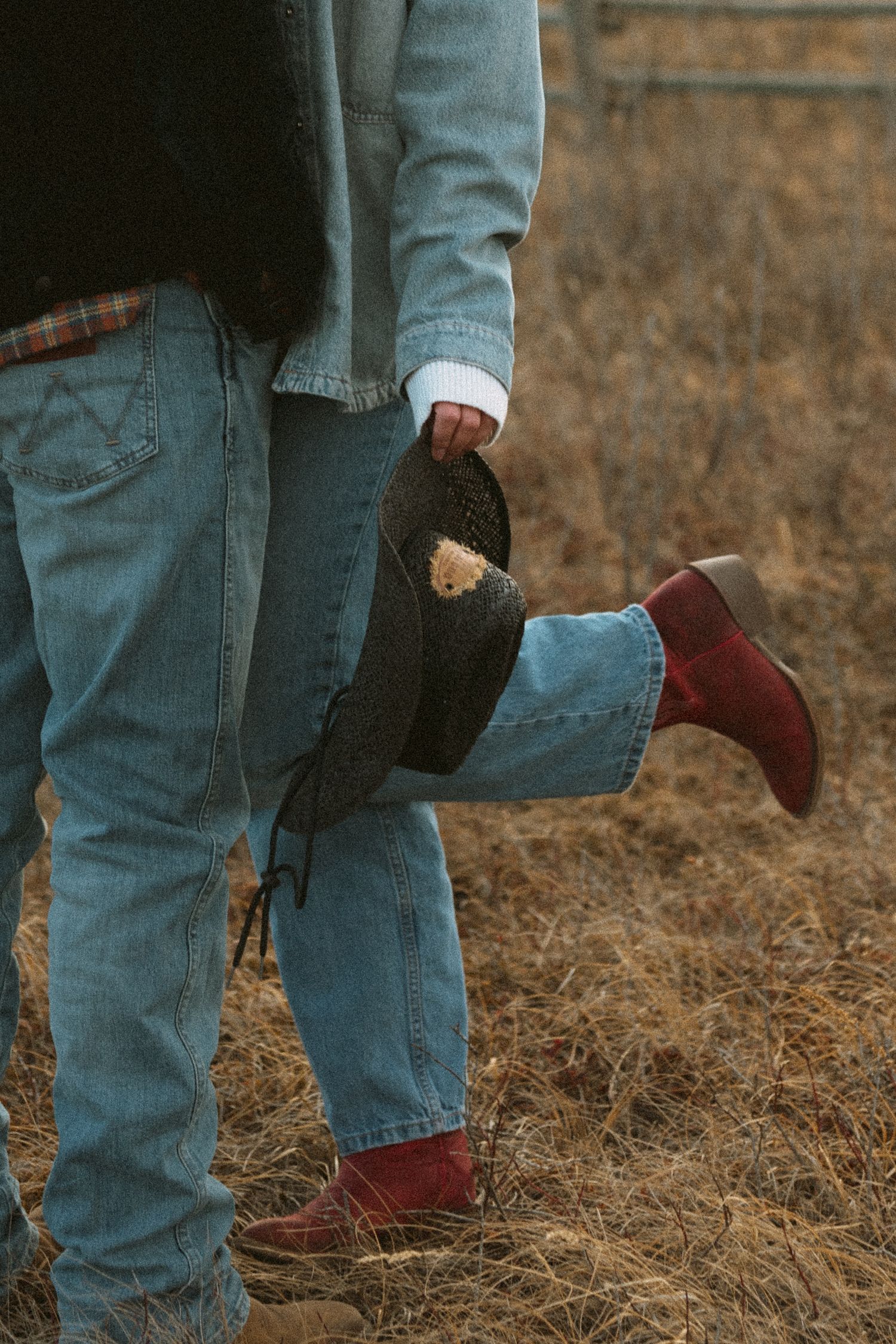 Close-up of denim jeans and burgundy boots against dry grass in an outdoor setting.